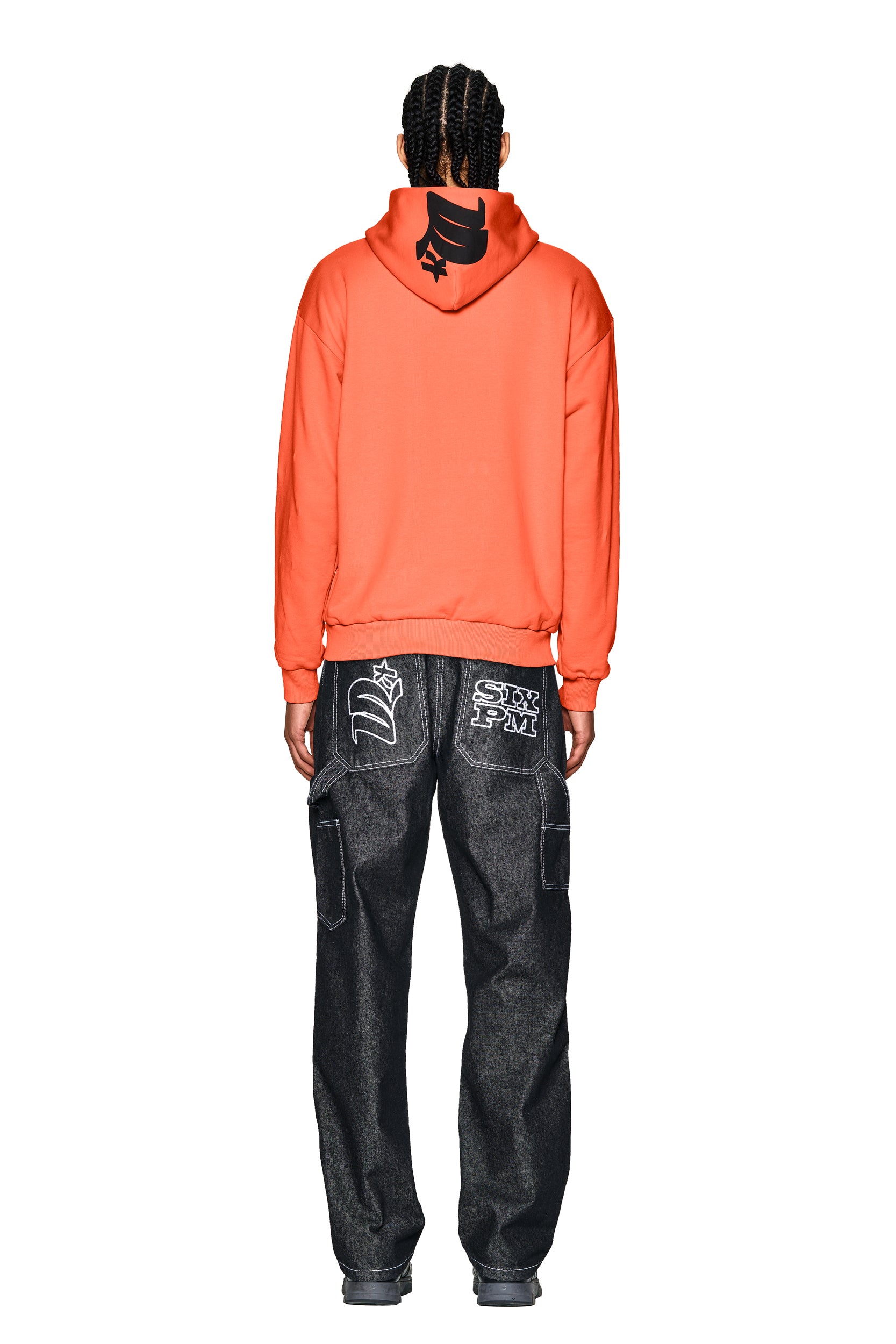 A person with braided hair faces away, wearing the 6PM LOGO ZIP HOODIE ORANGE with a black hood graphic and loose dark jeans featuring bold white graphics on the back pockets.