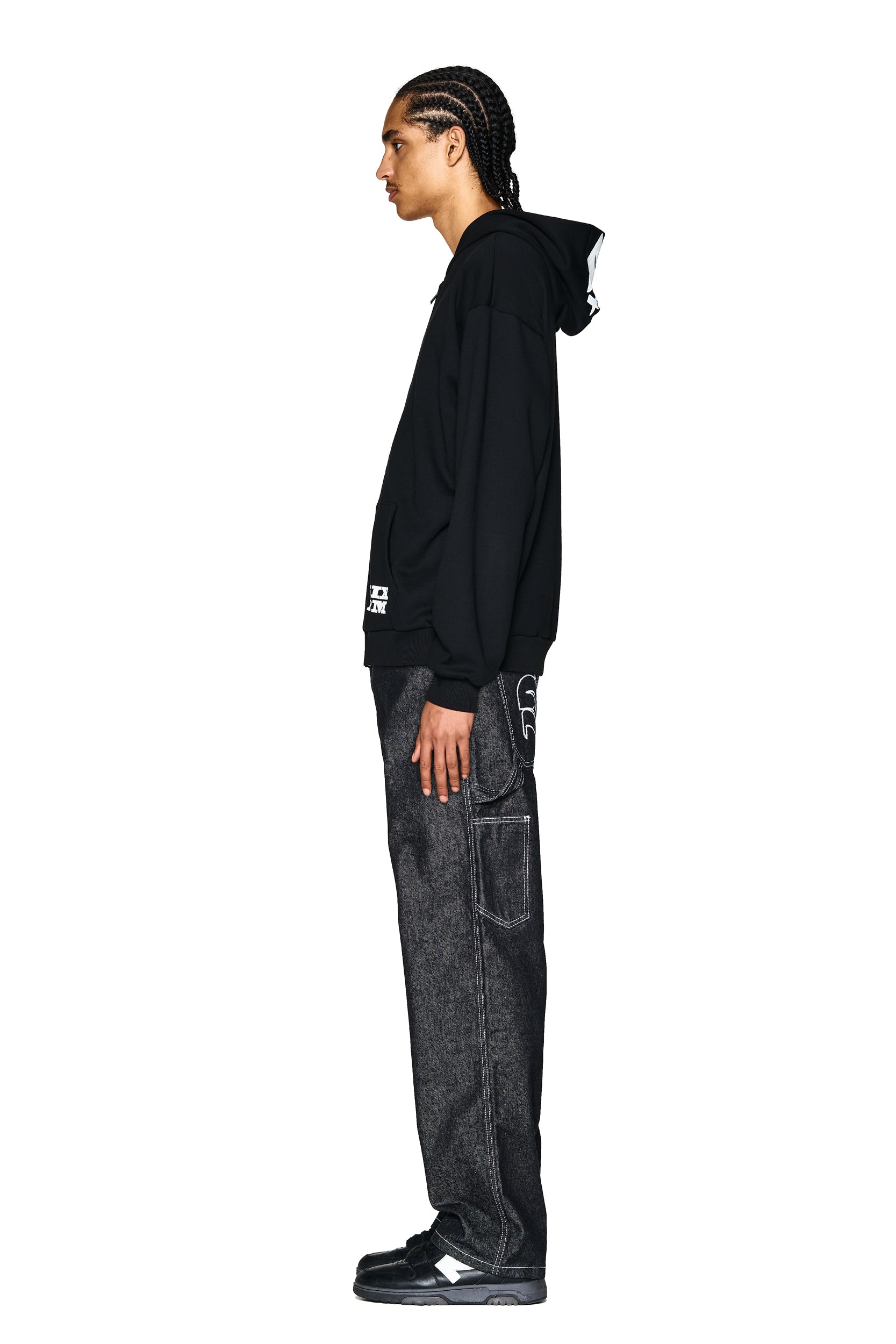 A person with braided hair stands in profile, wearing the 6PM LOGO ZIP HOODIE BLACK, dark loose-fitting jeans with visible stitching, and black sneakers, against a plain white background.