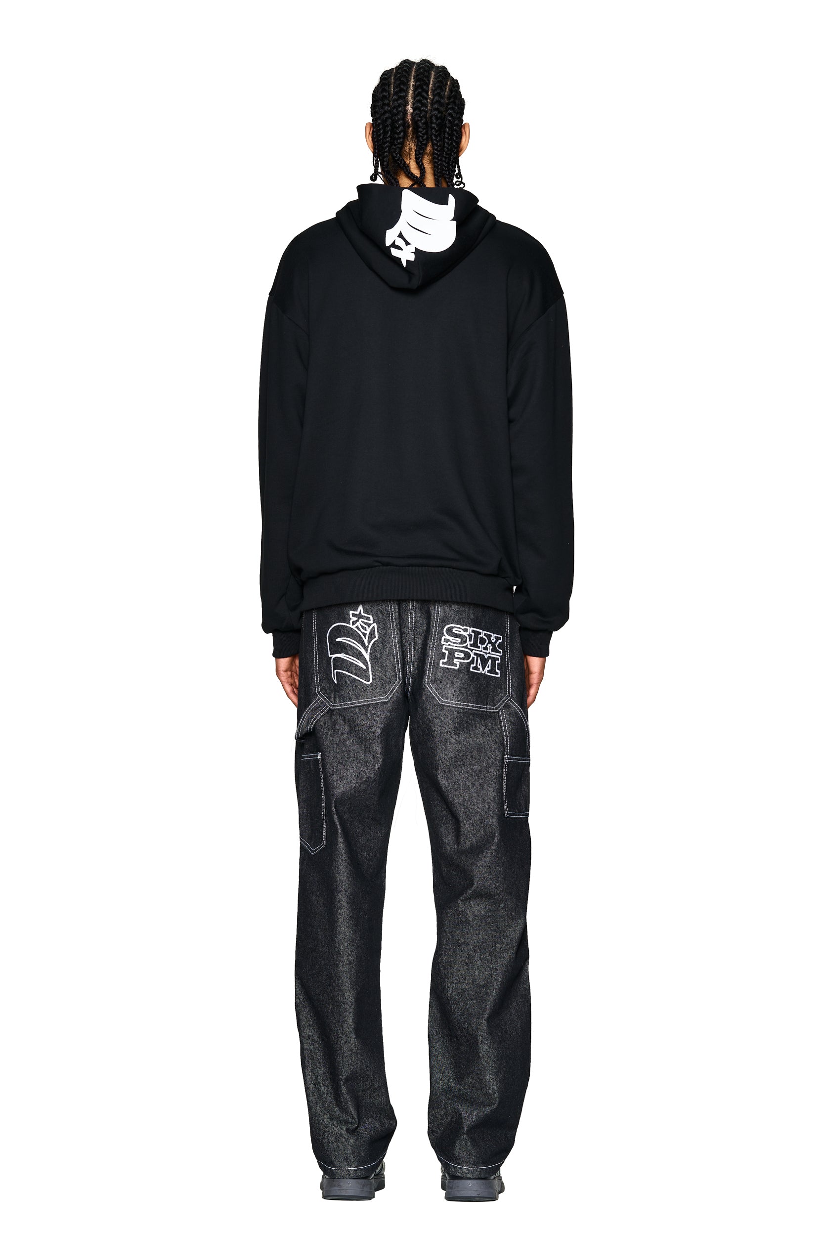 A person with braided hair faces away, wearing the 6PM LOGO ZIP HOODIE BLACK with a white hood graphic and dark denim jeans featuring large white graphics and "STAY PAW" text on the back pockets.