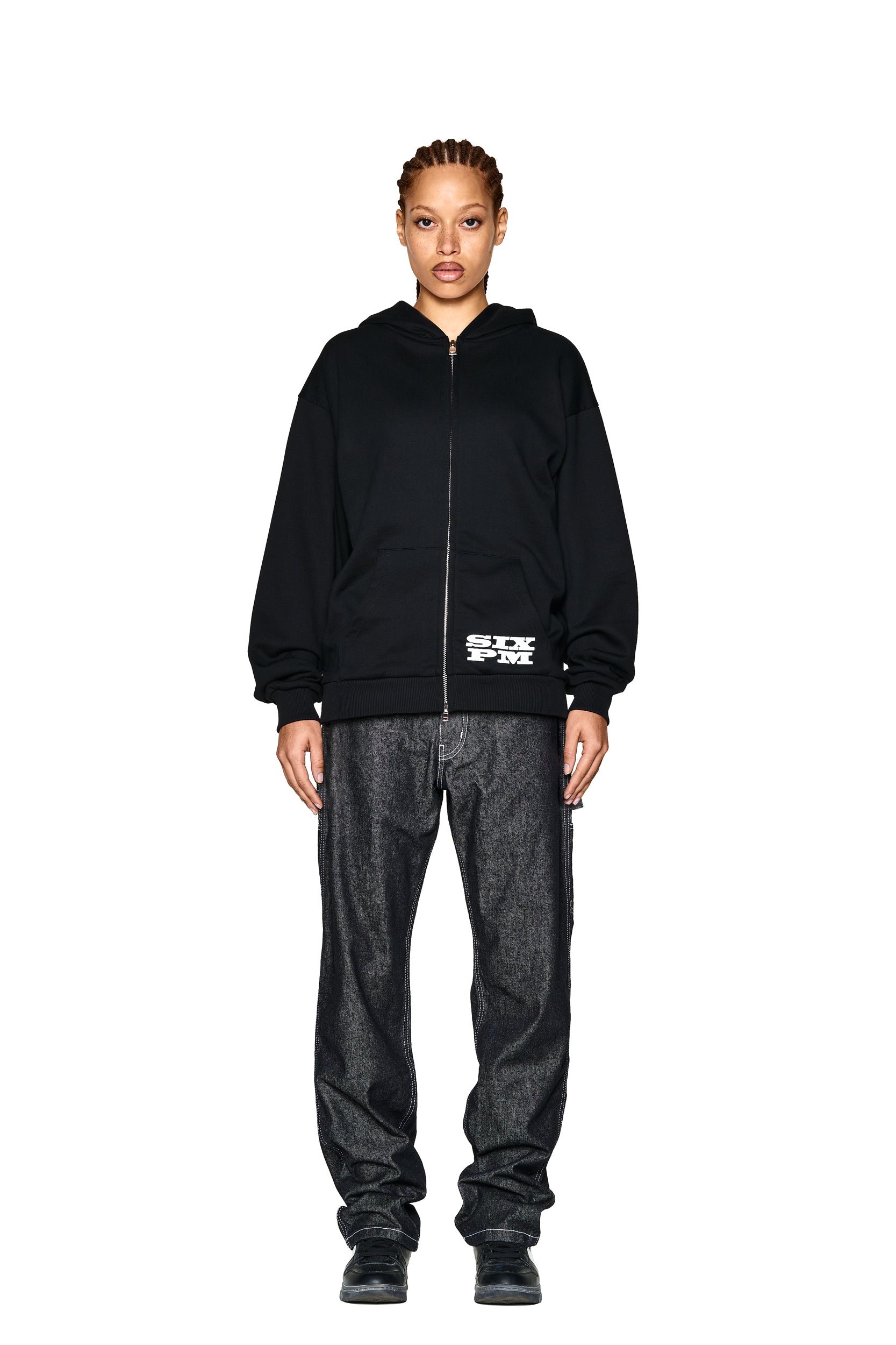 A person stands facing forward against a white background, wearing the 6PM LOGO ZIP HOODIE BLACK with a white graphic near the hem, paired with loose dark jeans and black shoes.