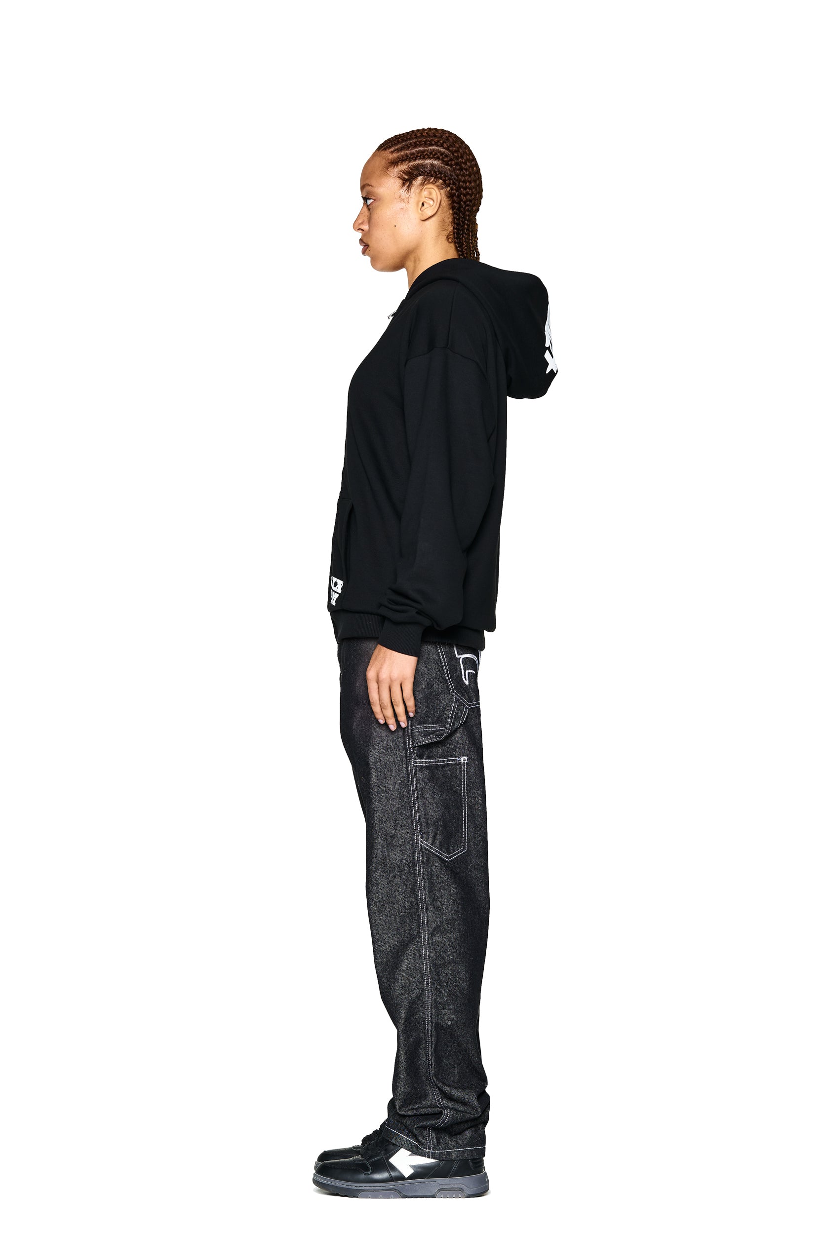 A person with braided hair stands in profile against a white background, wearing the 6PM LOGO ZIP HOODIE BLACK, dark jeans with white stitching, and black and white sneakers.