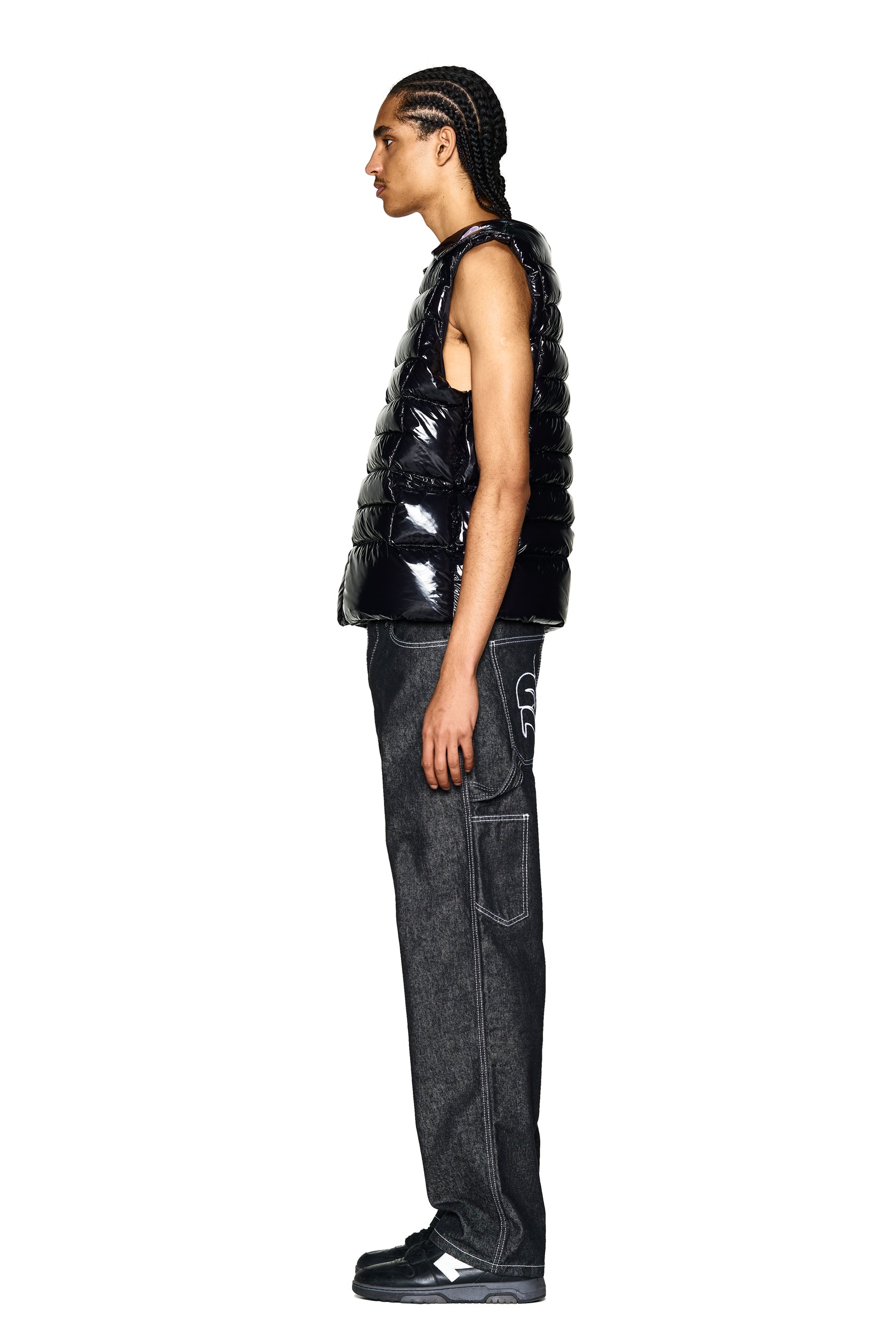 A person with braided hair stands in profile against a white background, wearing the 6PM BULLET PROOF VEST BLACK, dark wide-leg jeans with large pockets, and black sneakers.