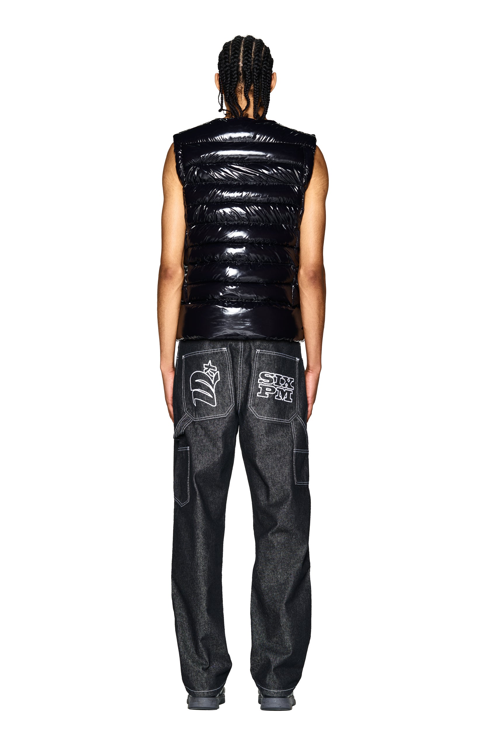 A person with braided hair faces away, wearing the 6PM BULLET PROOF VEST BLACK puffer and loose dark jeans featuring graphic patches on the back pockets, set against a plain white background.