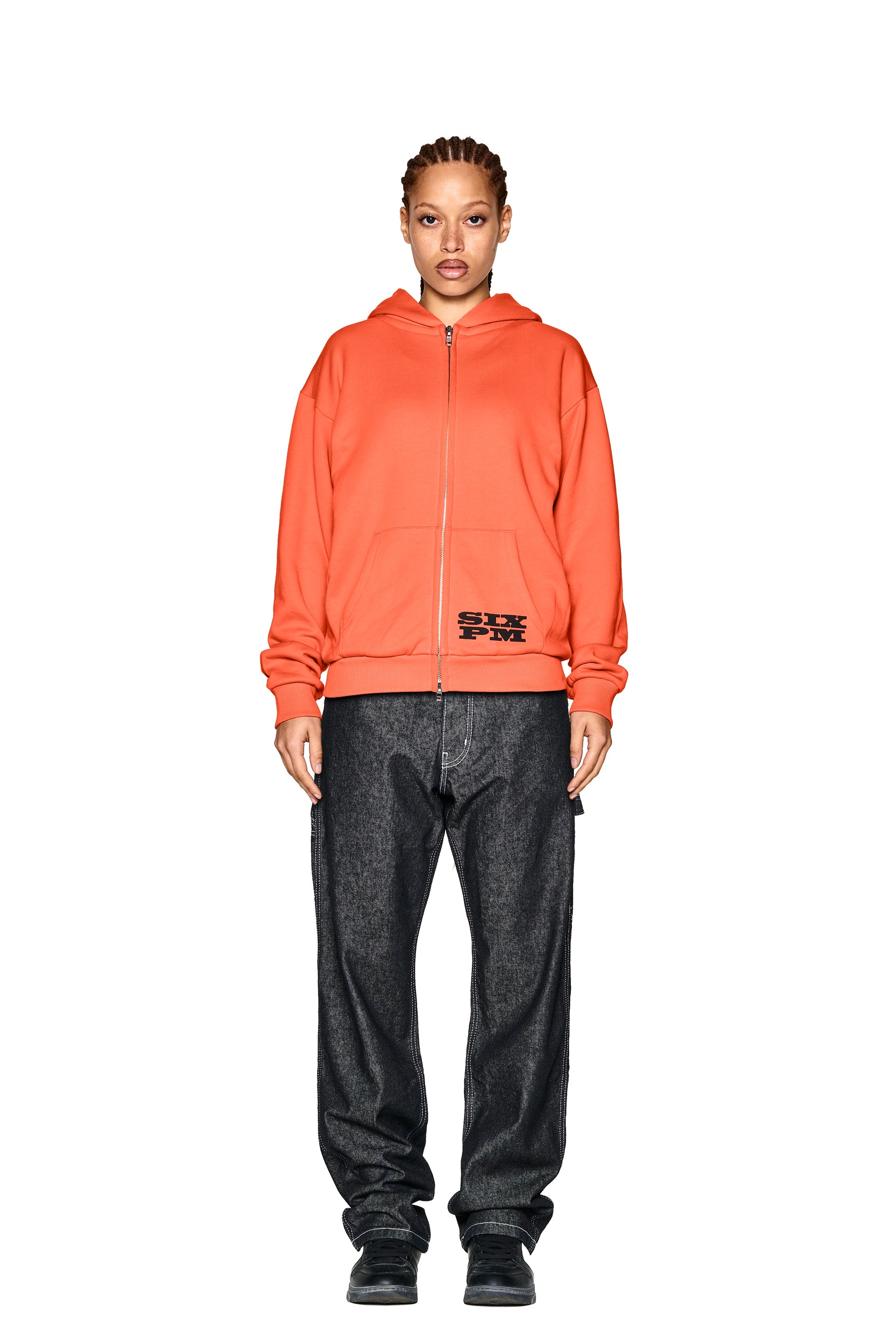 A person faces forward against a plain white background, wearing the 6PM LOGO ZIP HOODIE ORANGE paired with loose-fitting dark jeans and black shoes.