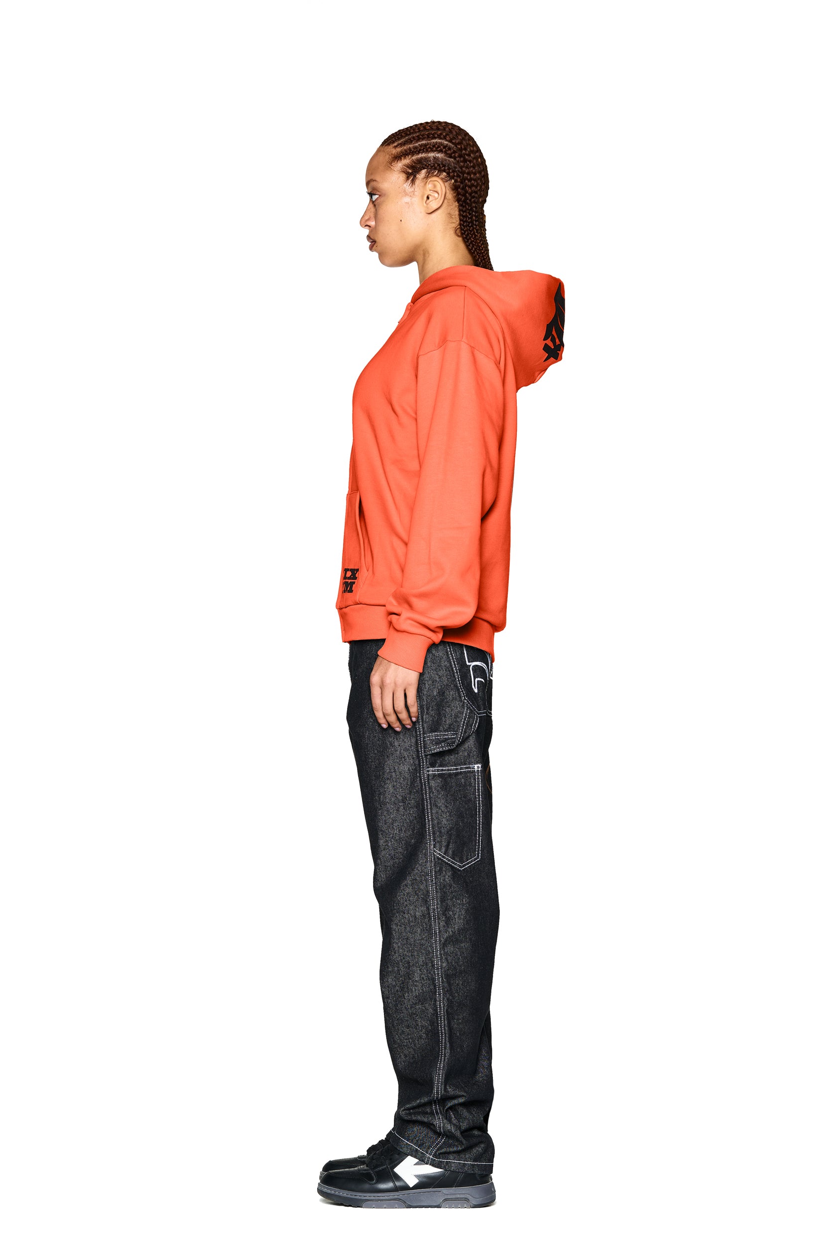 A person with braided hair stands in profile against a white background, wearing the 6PM LOGO ZIP HOODIE ORANGE, dark jeans with white stitching, and black and white sneakers.