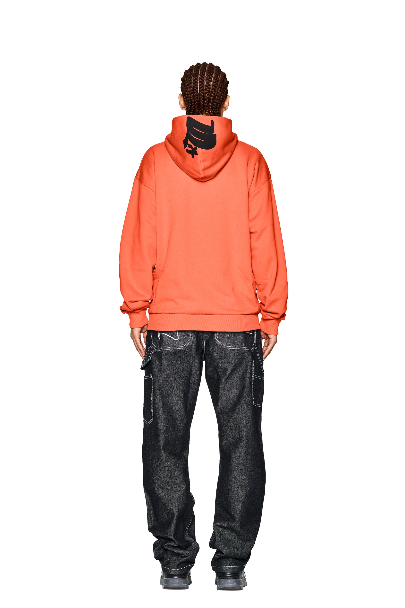 A person with braided hair faces away, wearing the 6PM LOGO ZIP HOODIE ORANGE with a black hood graphic, loose dark jeans with white stitching, and gray sneakers against a white background.