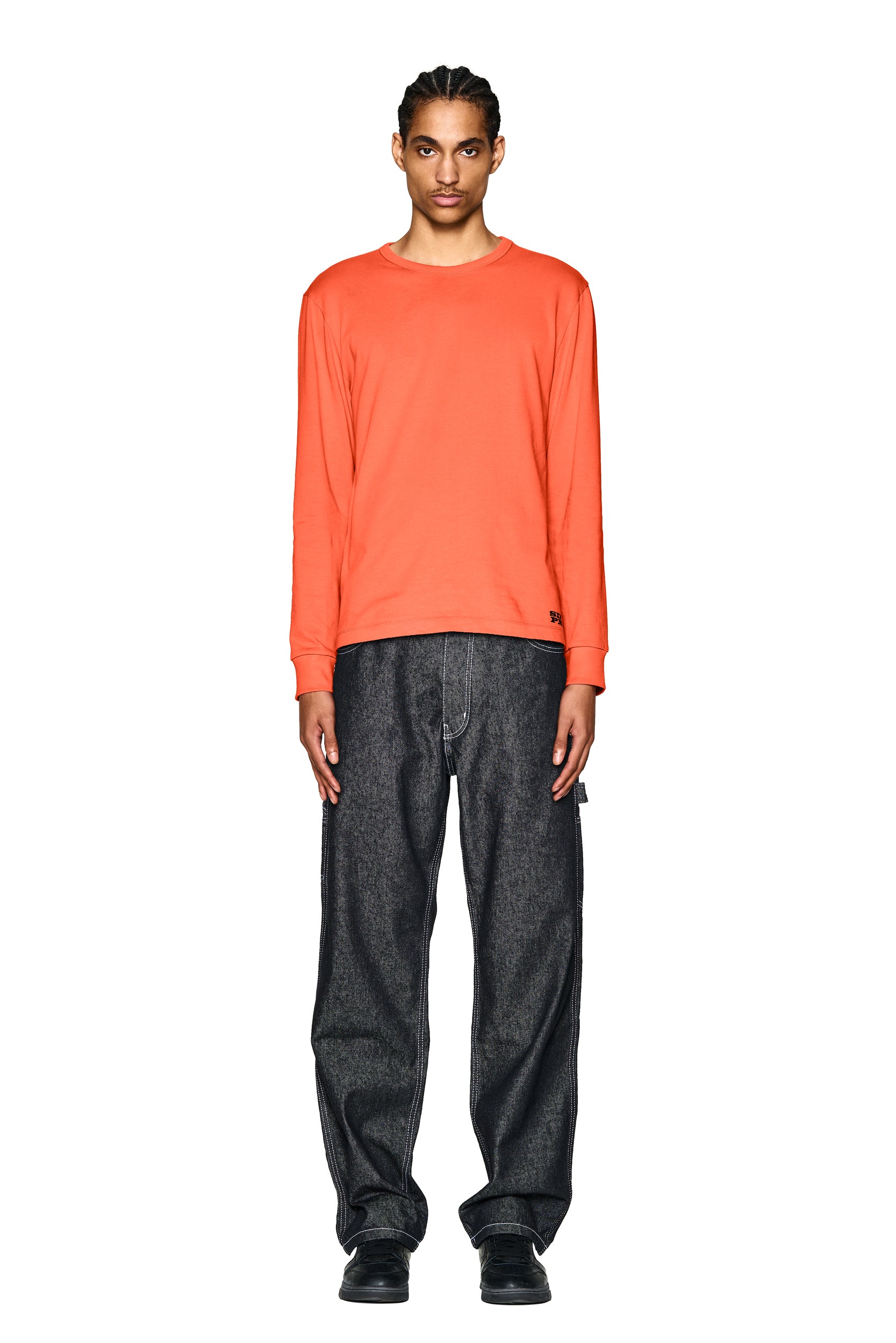 A person stands facing forward against a plain white background, wearing the 6PM DEVIL LONG SLEEVE ORANGE shirt, loose dark jeans, and black shoes.