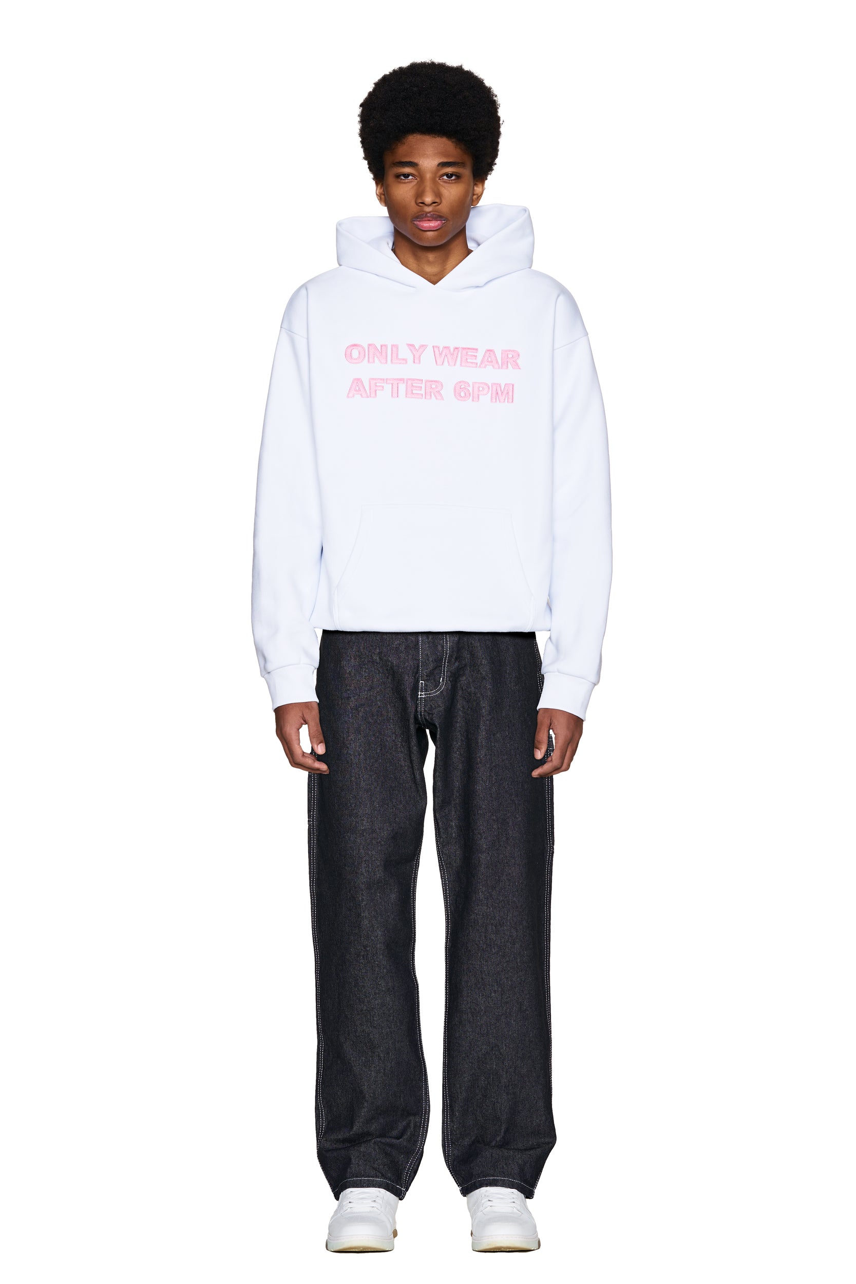 A young person with an afro wears the 6PM ONLY WEAR AFTER 6PM HOODIE 2.0 in white, styled with loose black jeans and white sneakers, standing against a plain white background.