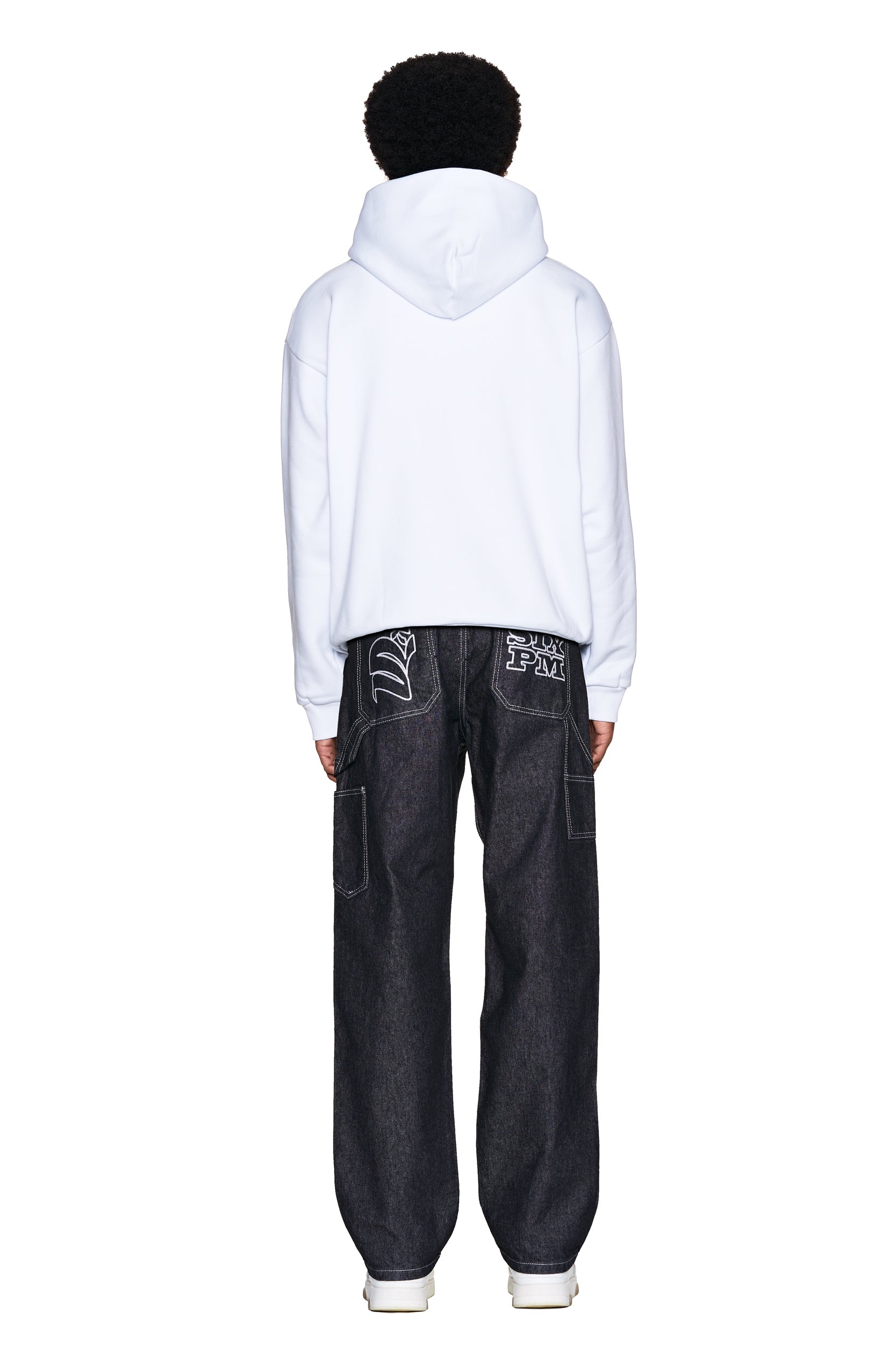 A person faces away, wearing the 6PM ONLY WEAR AFTER 6PM HOODIE 2.0 in white, paired with loose dark jeans featuring white graphic designs on the back pockets, set against a plain white background.