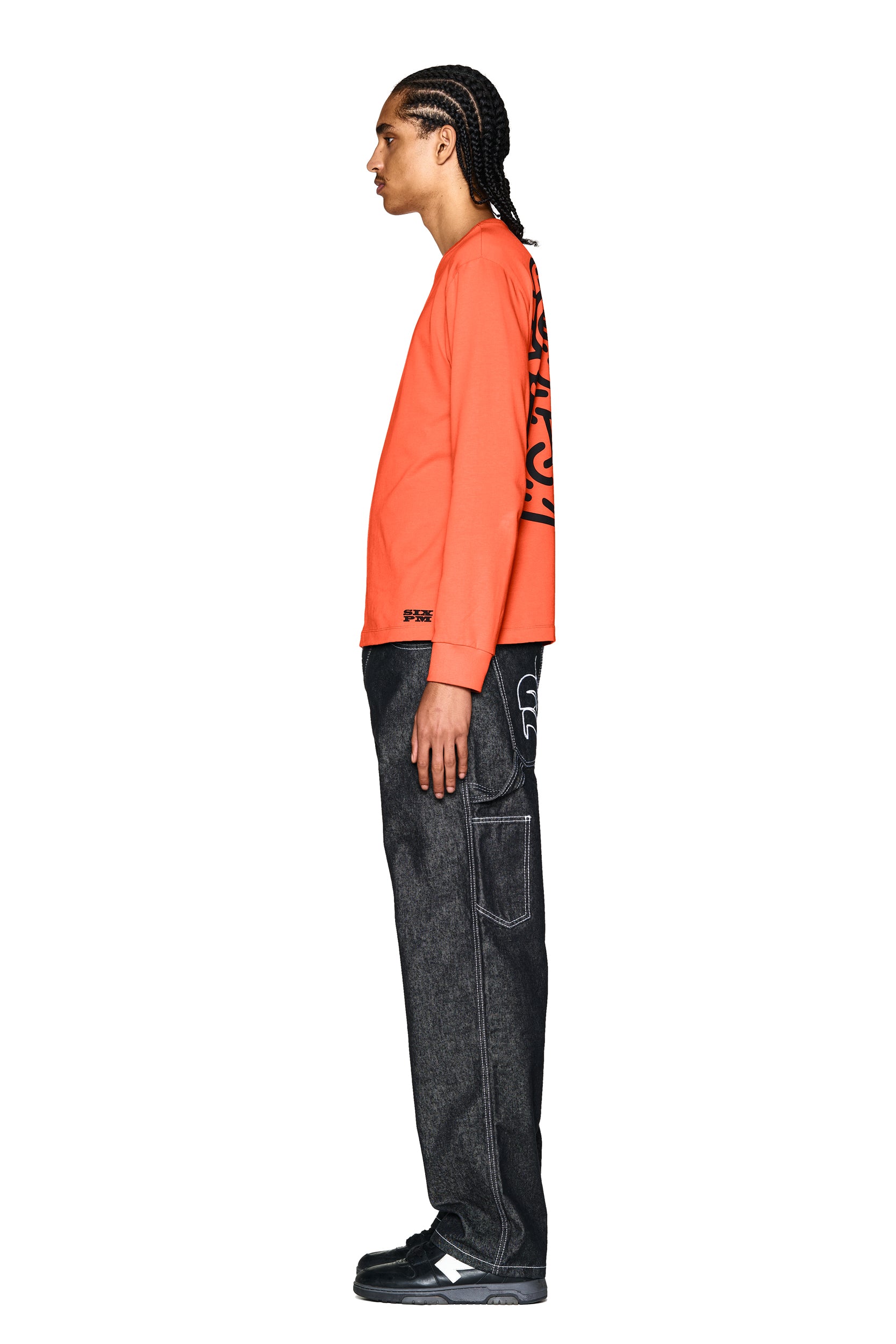 A person with braided hair stands in profile against a white background, wearing the 6PM DEVIL LONG SLEEVE ORANGE shirt, dark loose-fitting jeans with white stitching, and black sneakers.