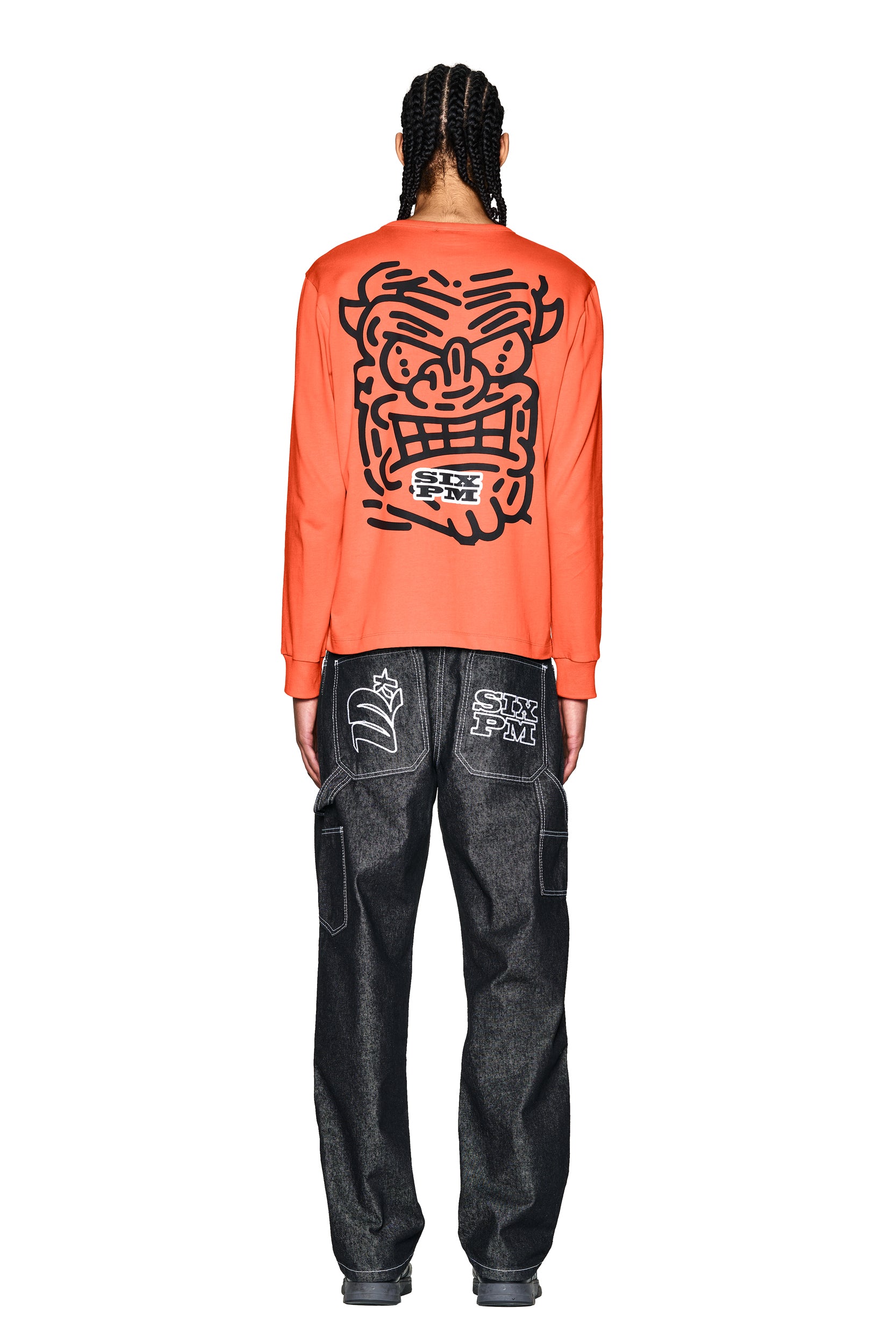 A person with braided hair faces away, wearing the 6PM DEVIL LONG SLEEVE ORANGE shirt featuring a bold cartoon face graphic on the back, paired with dark jeans with white graphic patches on the rear pockets. The background is white.