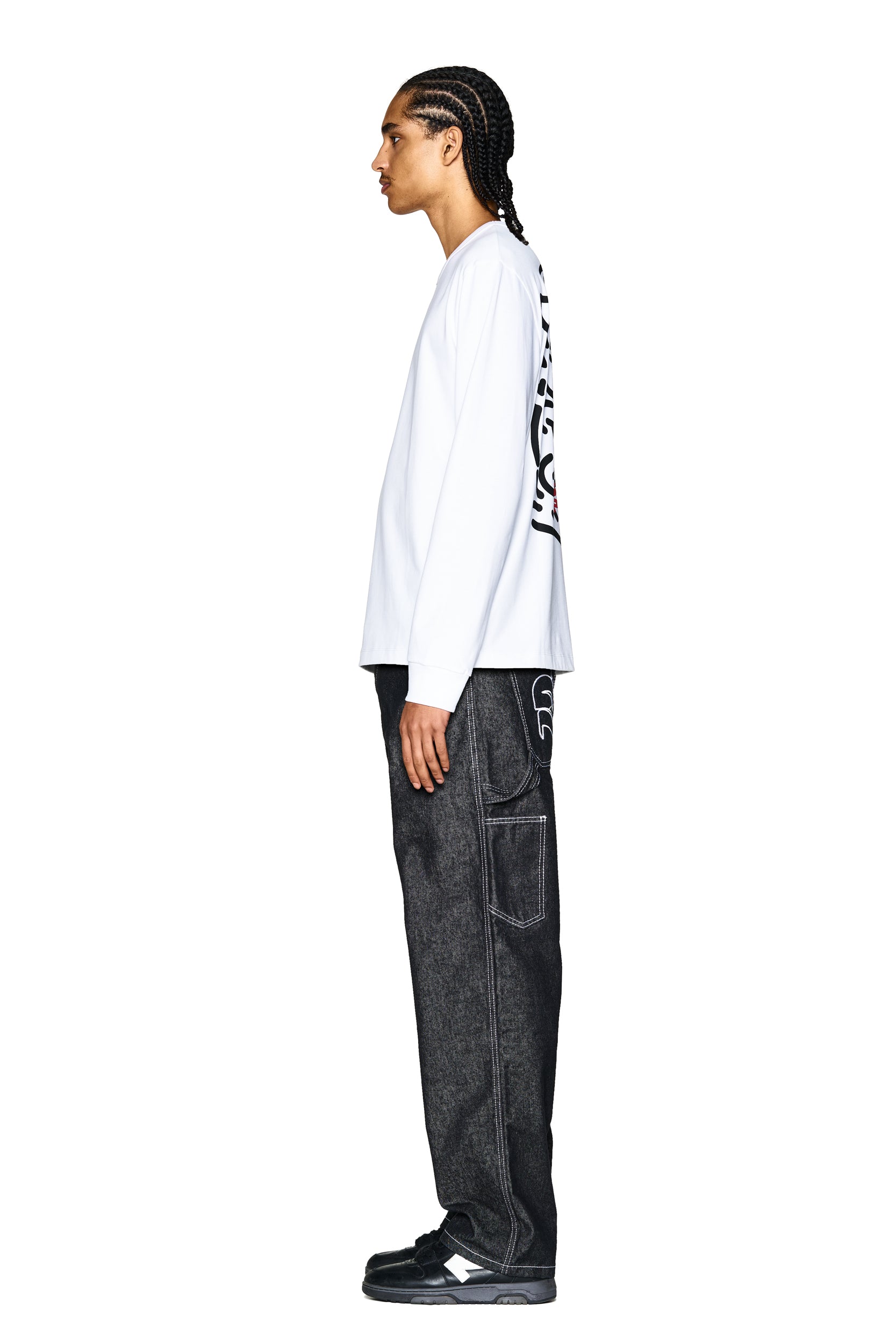 A person with braided hair stands in profile against a plain white background, wearing the 6PM DEVIL LONG SLEEVE WHITE shirt featuring black back graphics, paired with dark loose-fitting jeans with white stitching and black sneakers.