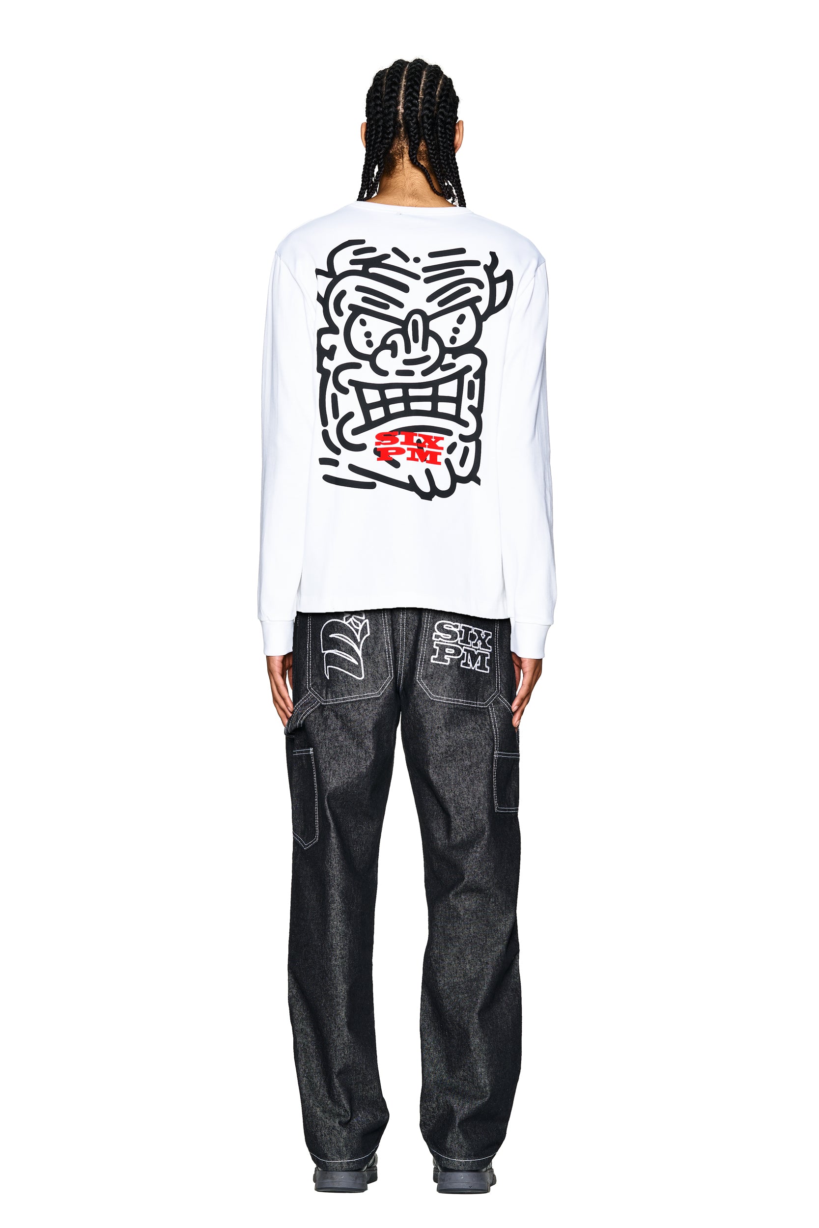 A person with braided hair faces away, wearing the 6PM DEVIL LONG SLEEVE WHITE shirt with a bold cartoon face and red back detail, paired with loose dark denim jeans featuring patched back pockets.