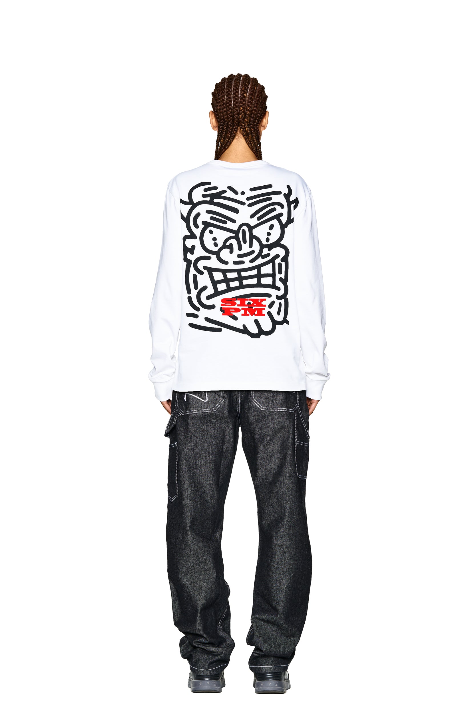A person with braided hair, seen from behind, wears the 6PM DEVIL LONG SLEEVE WHITE featuring a bold black cartoon face and red text on the back, styled with loose dark jeans and gray sneakers.