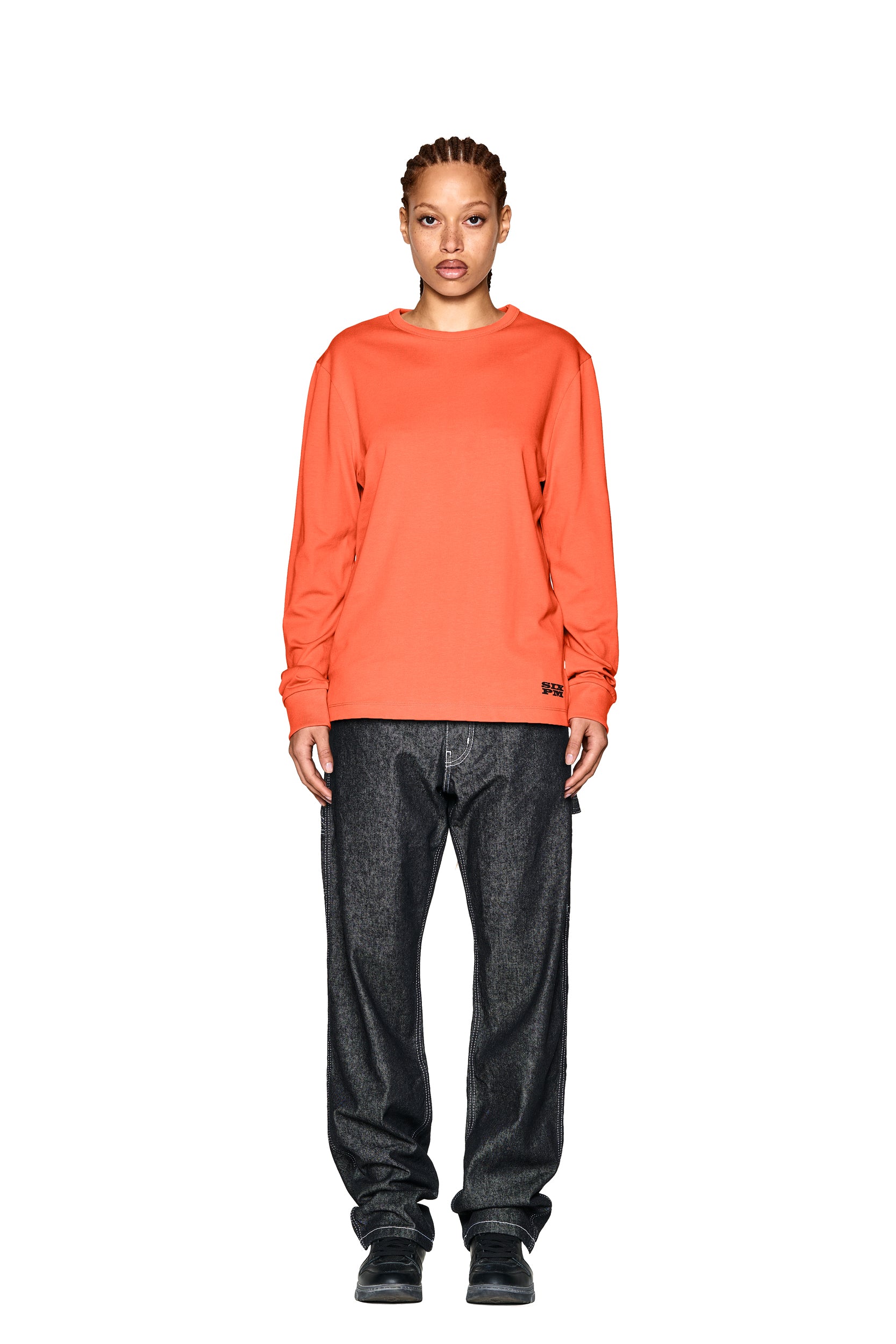 A person stands facing forward against a plain white background, wearing the 6PM DEVIL LONG SLEEVE ORANGE shirt, loose-fitting dark jeans, and black shoes.