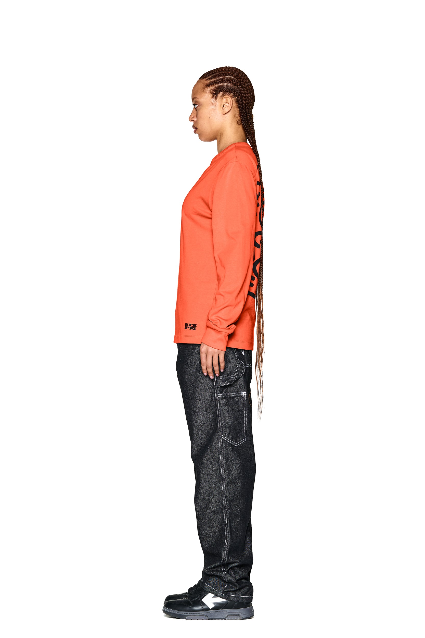 A person with long braided hair stands in profile against a white background, wearing the 6PM DEVIL LONG SLEEVE ORANGE shirt with black sleeve text, dark jeans with large pockets, and black sneakers with white accents.