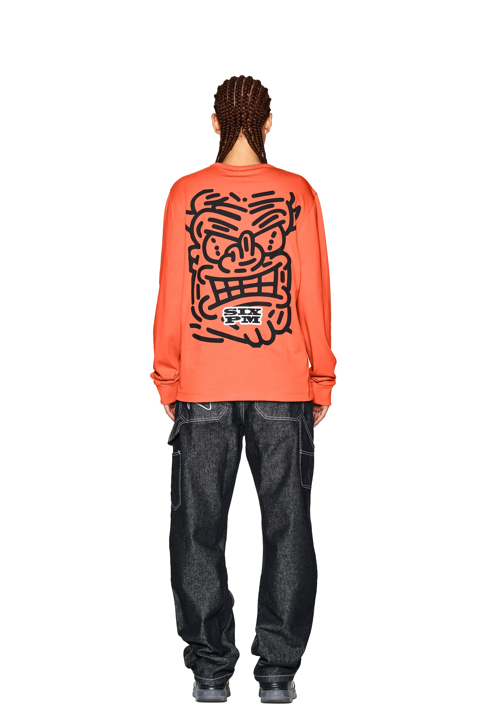 A person with braided hair is seen from behind wearing the 6PM DEVIL LONG SLEEVE ORANGE shirt, featuring a large black cartoon face graphic on the back, paired with dark loose-fitting jeans and gray shoes.