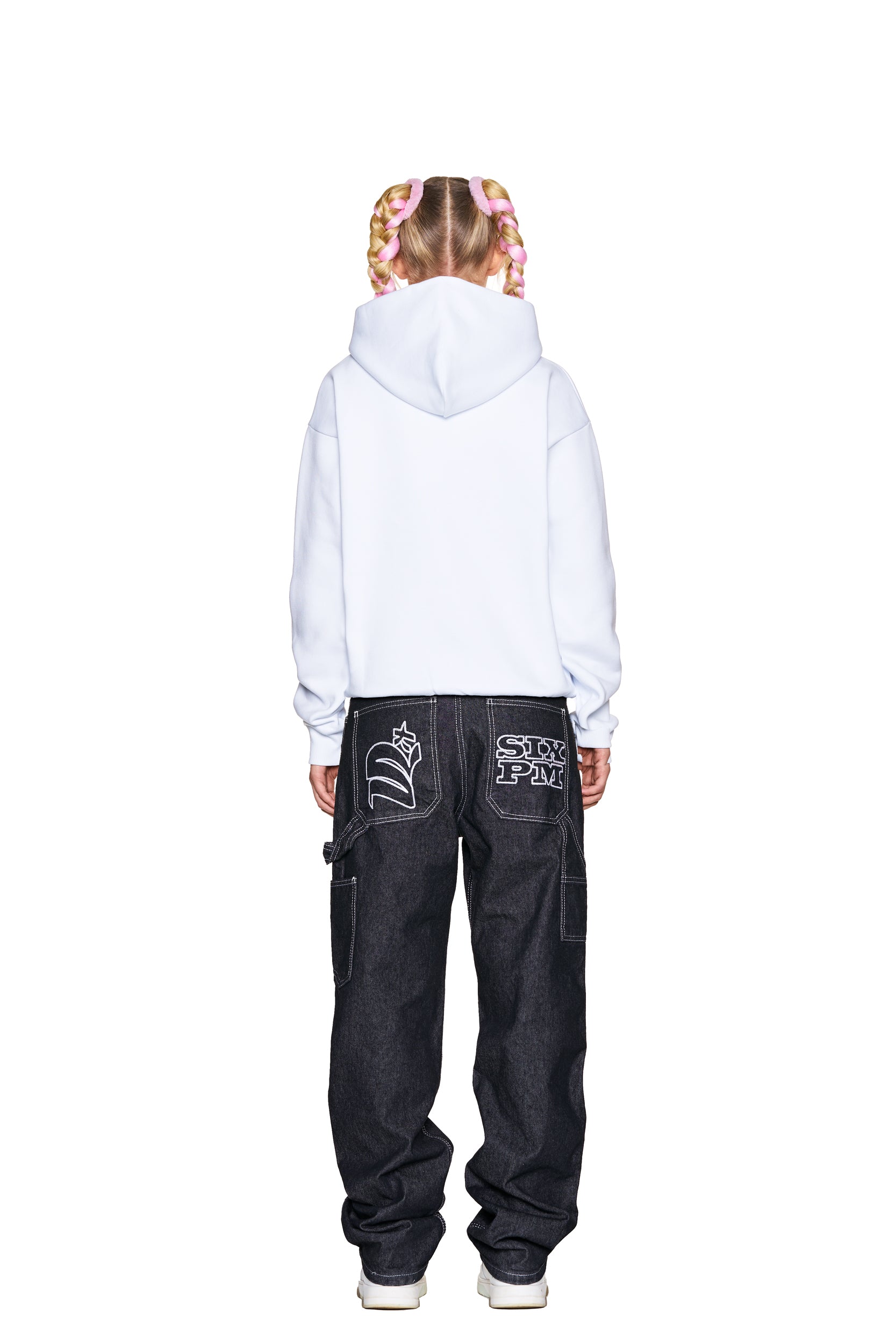 A person with pink braided hair faces away, wearing the 6PM ONLY WEAR AFTER 6PM HOODIE 2.0 WHITE and loose dark jeans with white graphic designs on the back pockets, set against a plain white background.