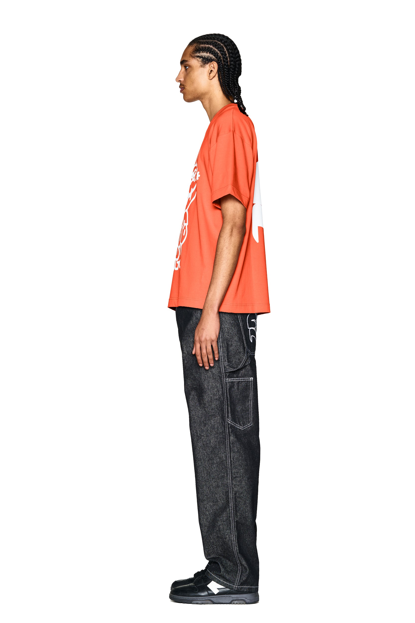 A person with braided hair stands in profile against a white background, wearing the 6PM DEMON T-SHIRT ORANGE, loose black denim jeans with contrast stitching, and black sneakers.