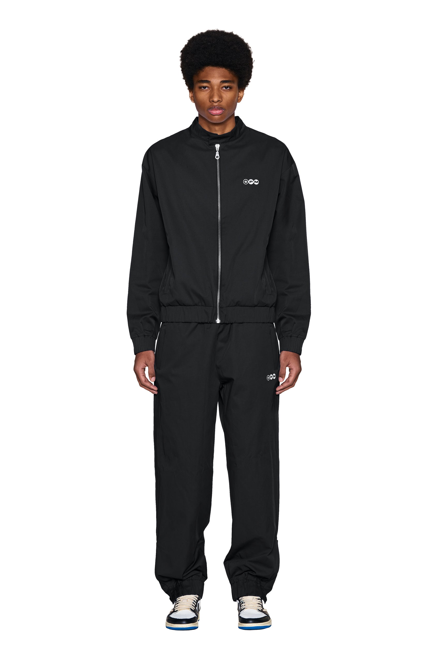 A person stands facing forward, wearing the 6PM TRACK JACKET BLACK and matching pants, both featuring small white logos on the chest and thigh. They pair the outfit with white sneakers against a plain white background.