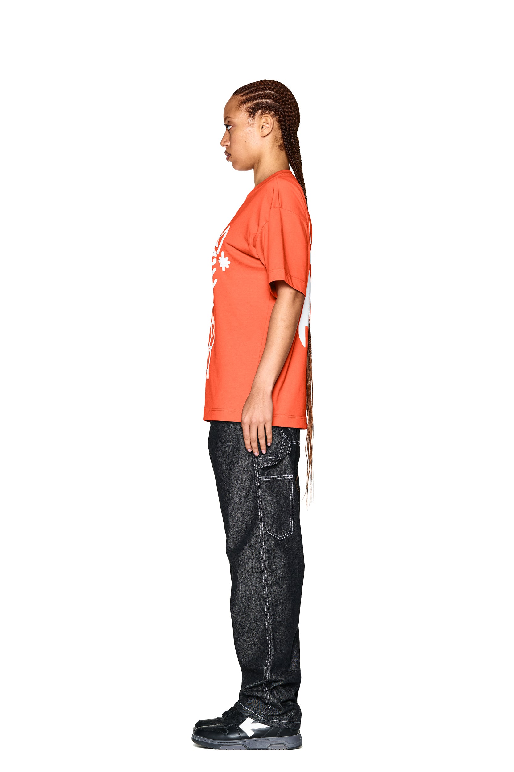 A person with long braided hair stands in profile against a white background, wearing the 6PM DEMON T-SHIRT ORANGE, dark jeans, and black sneakers.