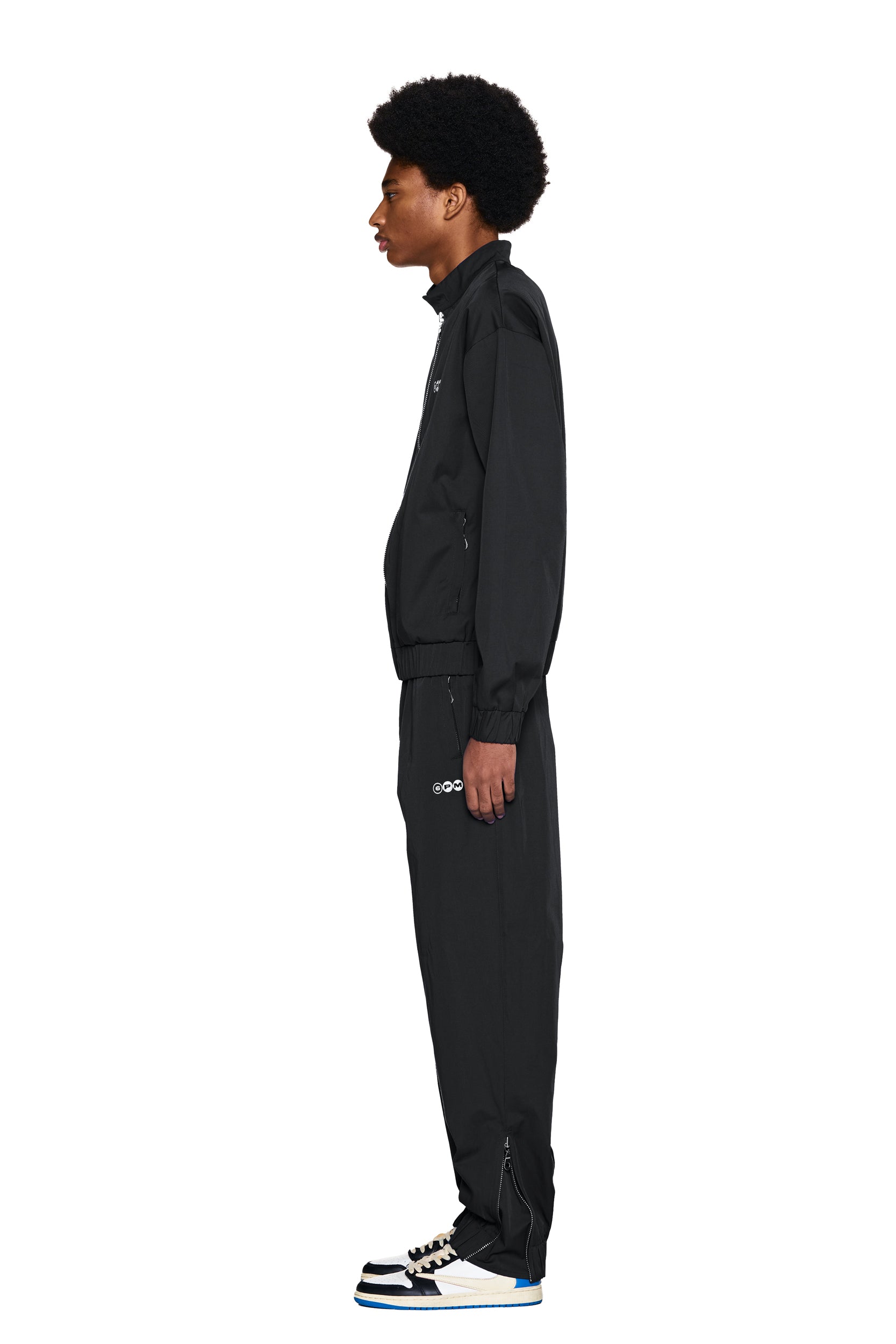 A person with an afro stands in profile, wearing the 6PM TRACK JACKET BLACK and black-and-white sneakers, against a plain white background.