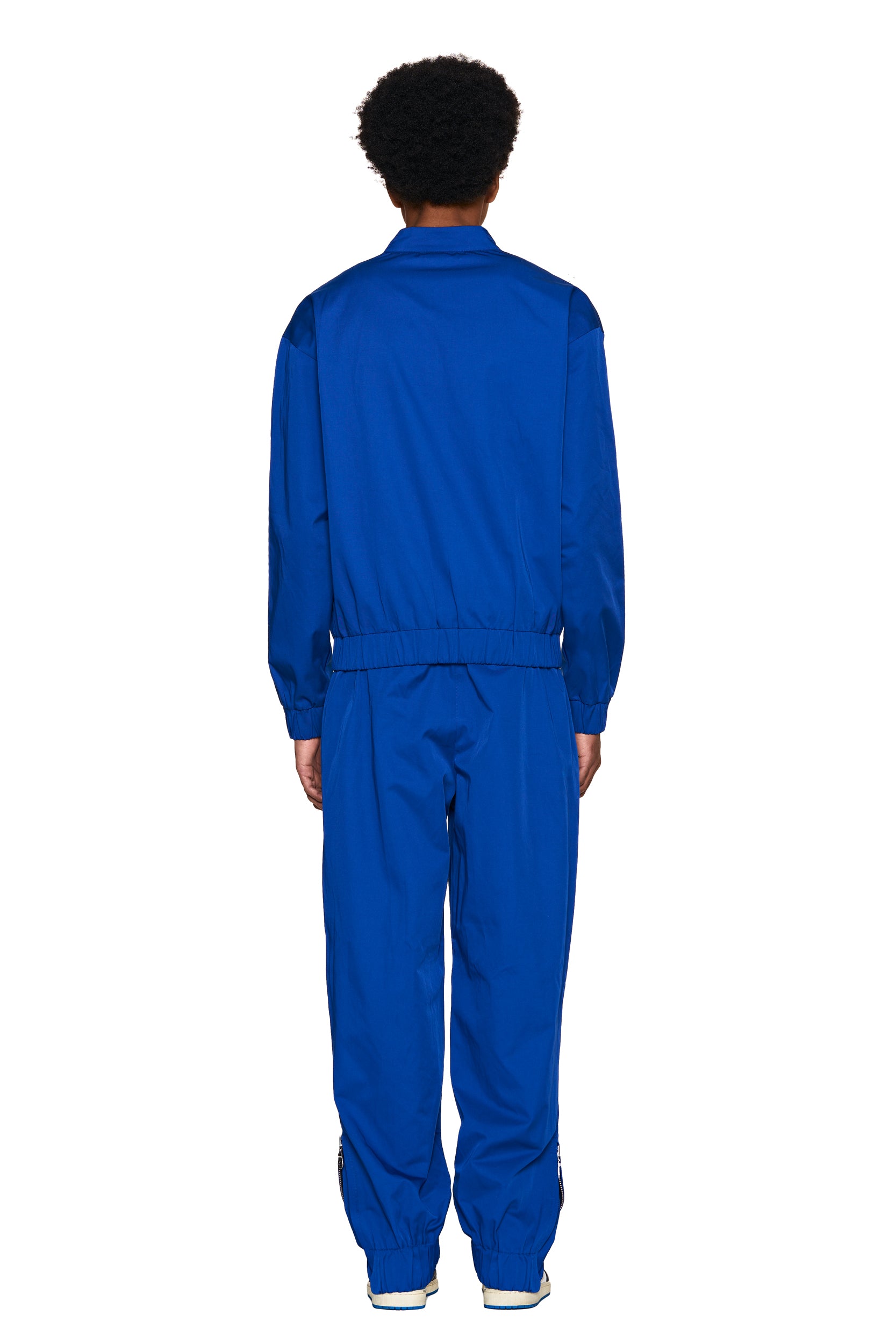 A person with short curly hair faces away, wearing the 6PM TRACK JACKET BLUE and white sneakers against a plain white background.