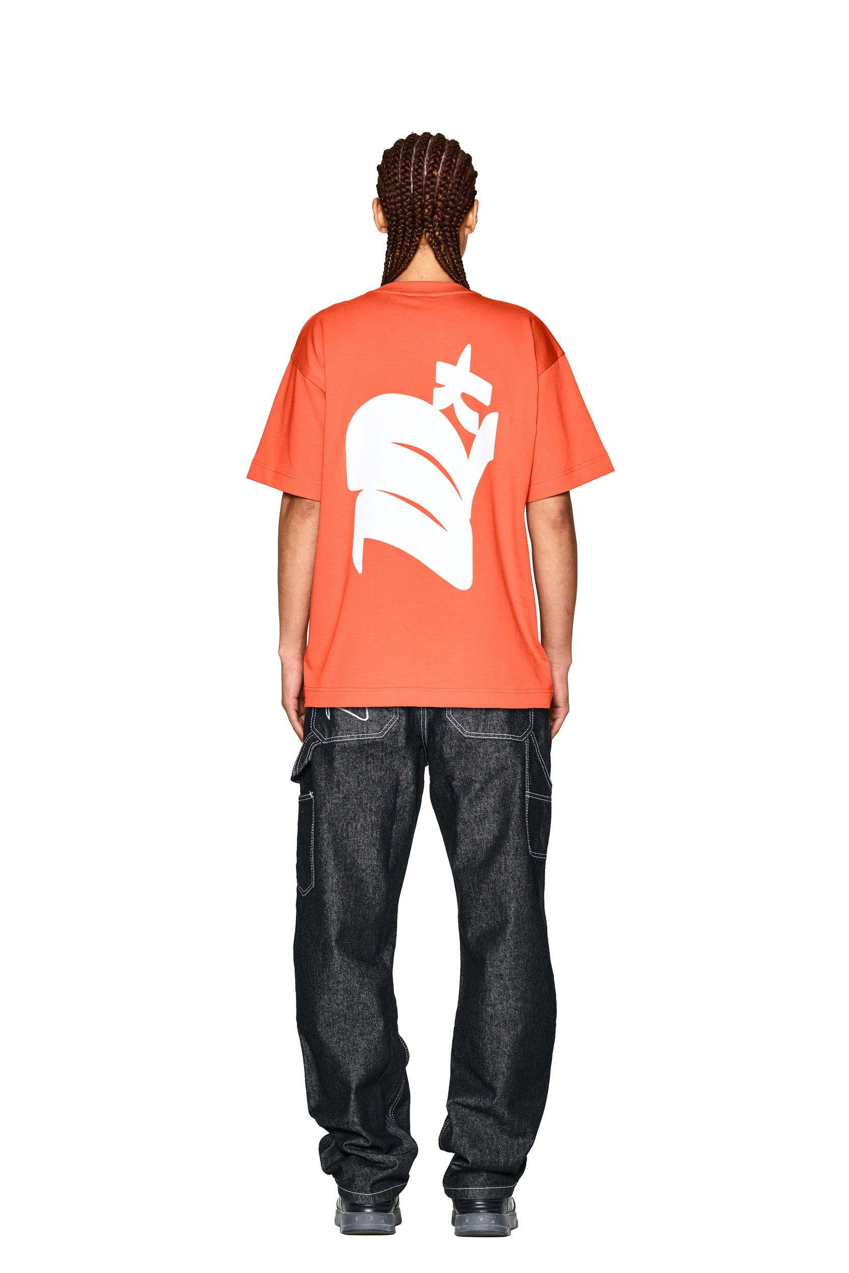 A person with braided hair wears the 6PM DEMON T-SHIRT ORANGE featuring a bold white abstract back graphic, paired with dark baggy jeans and gray sneakers, standing against a plain white background.