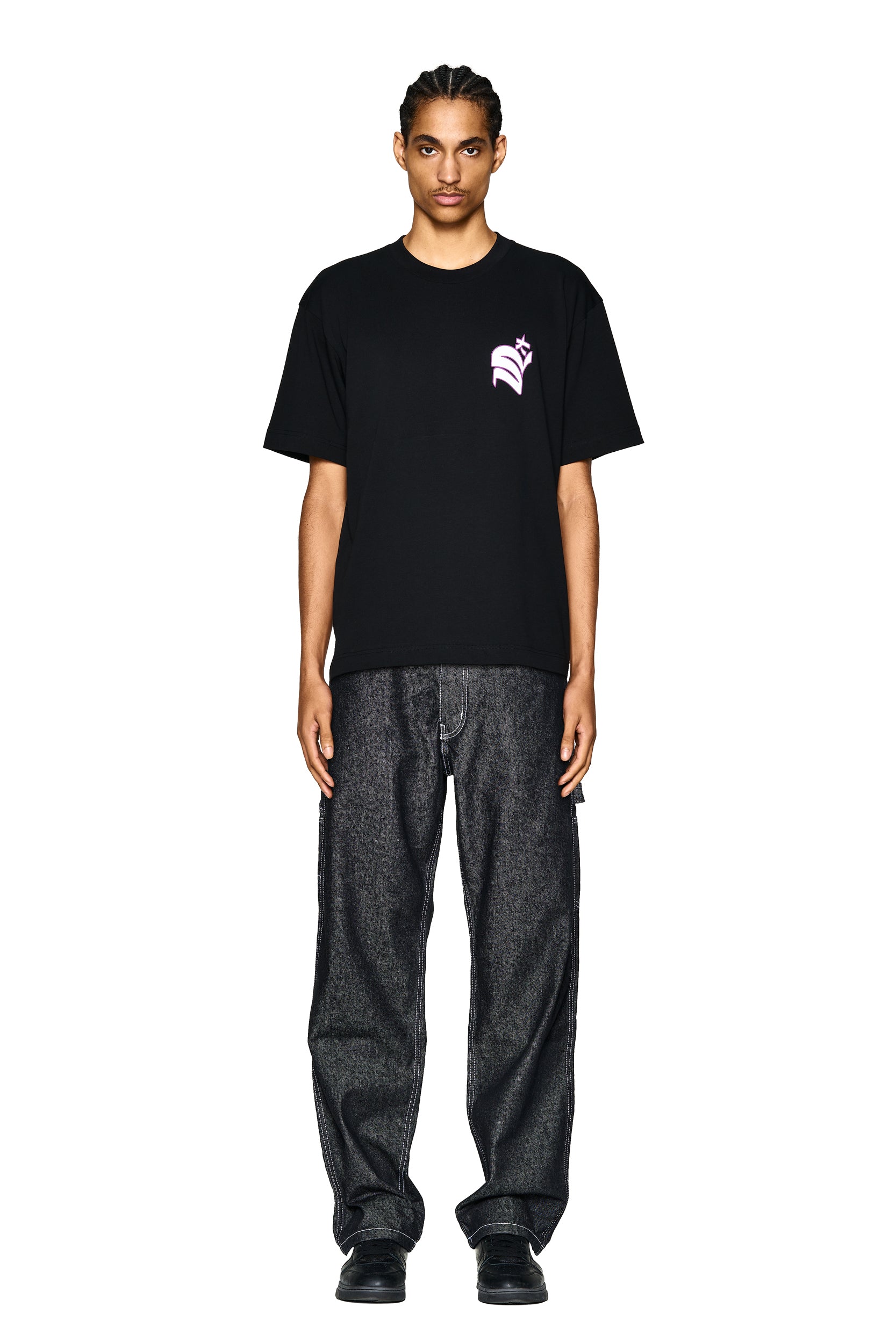 A person faces forward against a plain white background, wearing the 6PM LOGO T-SHIRT BLACK with a small pink chest graphic, loose dark jeans, and black shoes.