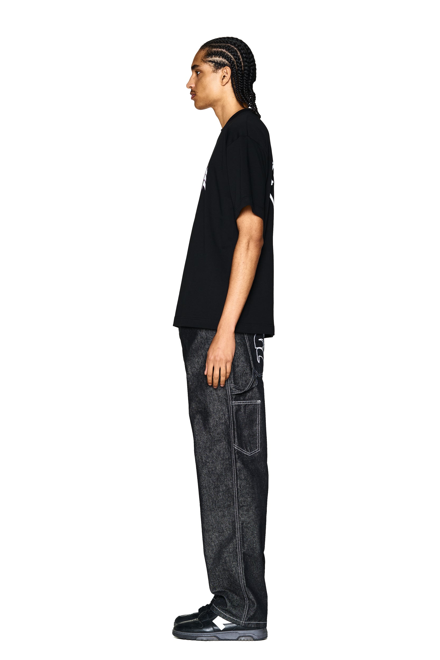 A person with braided hair stands in profile against a white background, wearing the 6PM LOGO T-SHIRT BLACK, loose dark jeans with white stitching, and black sneakers.