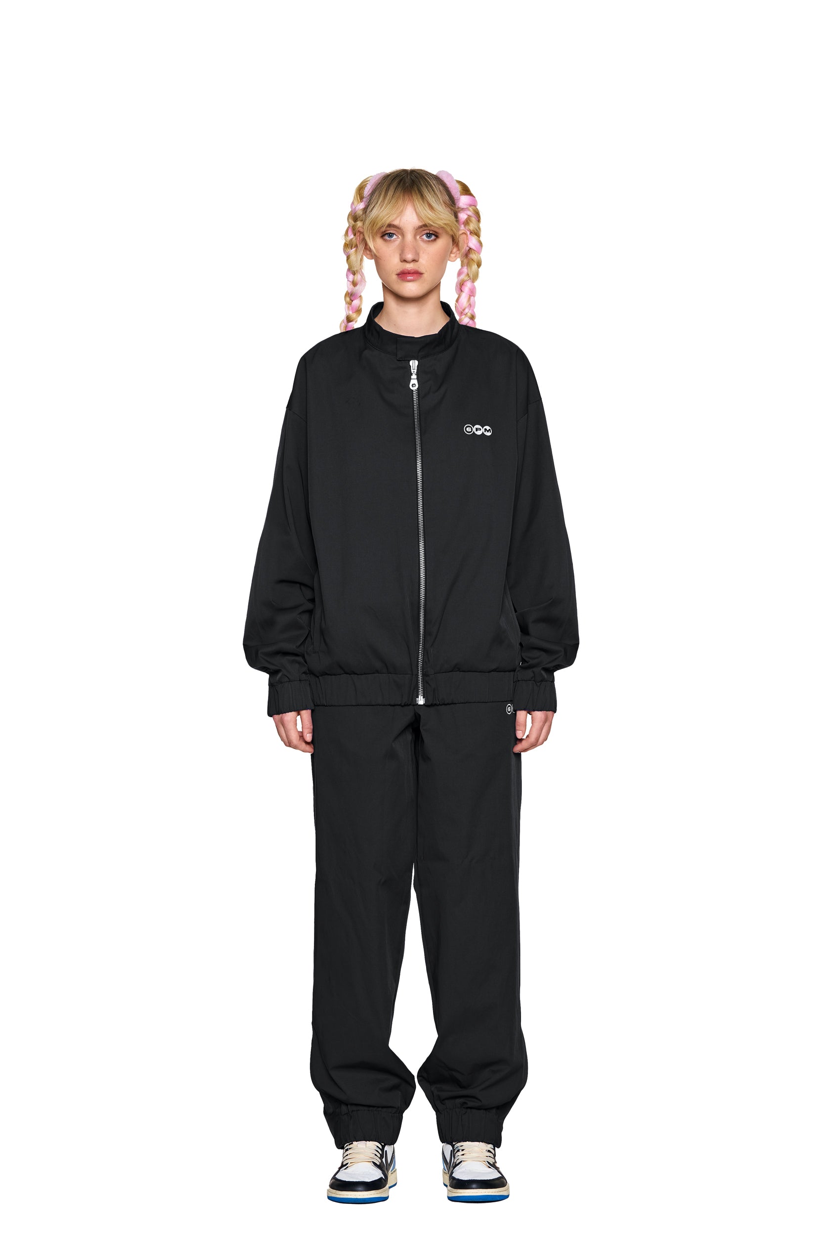A person with pink braided hair stands facing forward in a loose-fitting black zip-up jacket and 6PM TRACK PANTS BLACK, paired with white sneakers, against a plain white background.