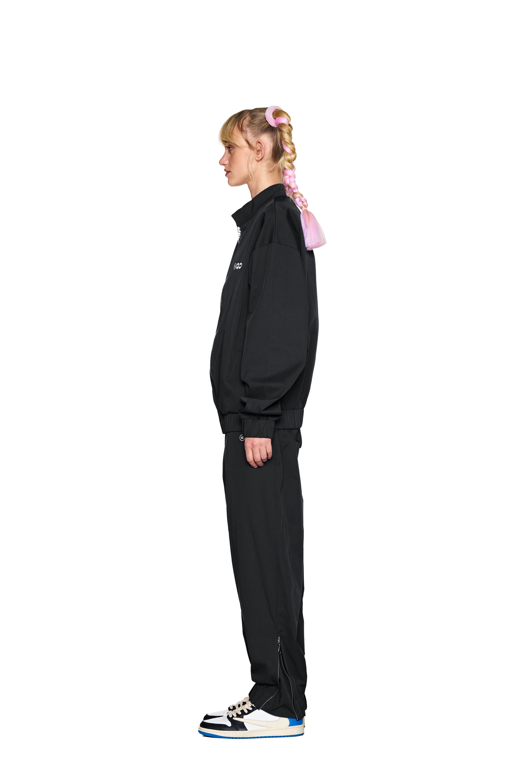 A person with long, pink braided hair stands in profile wearing the 6PM TRACK JACKET BLACK, matching black pants, and black-and-white sneakers with blue soles against a plain white background.