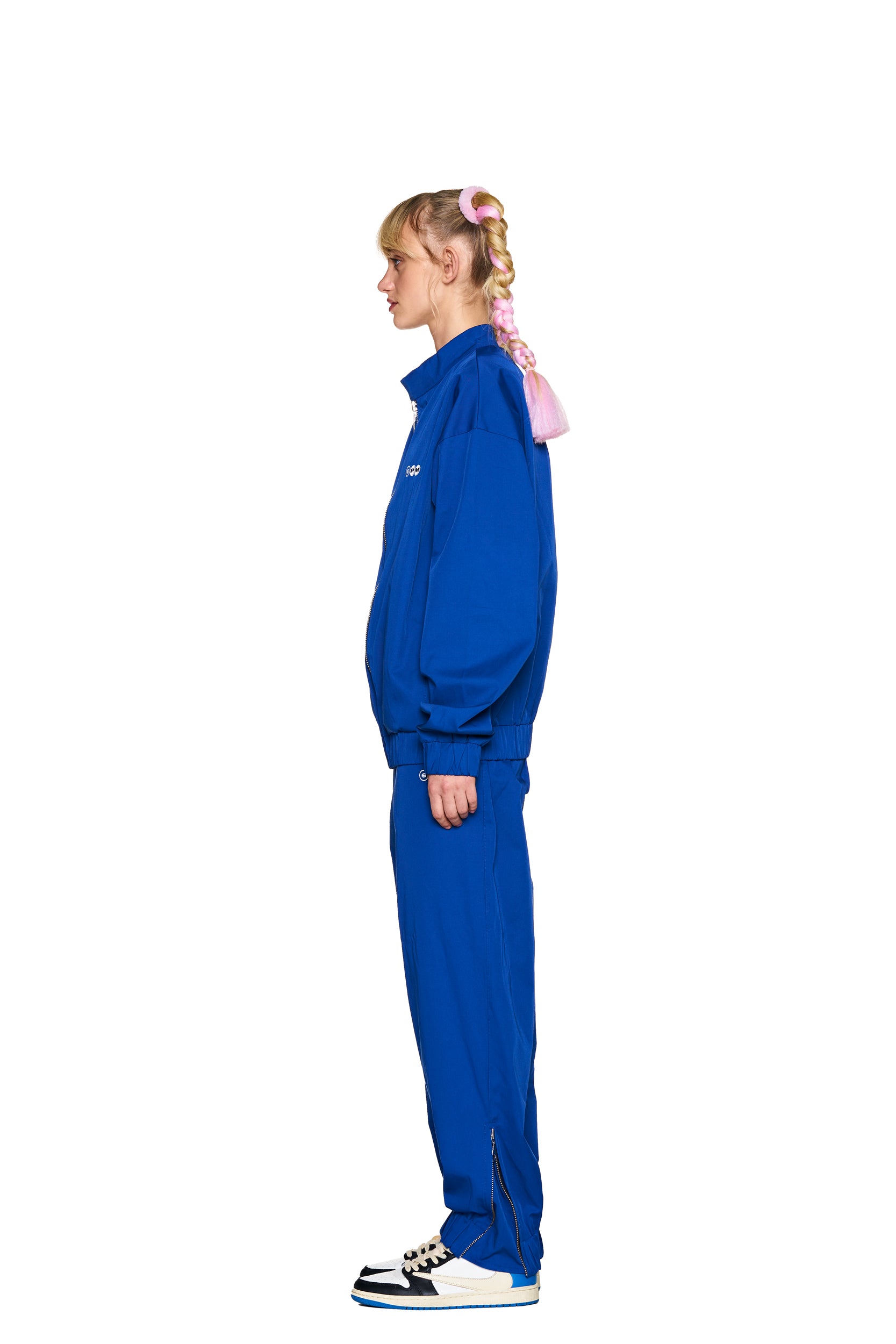 A person in profile stands against a white background, wearing 6PM TRACK PANTS BLUE and black-and-white sneakers. Their hair is in a braided ponytail with pink ribbon accents.