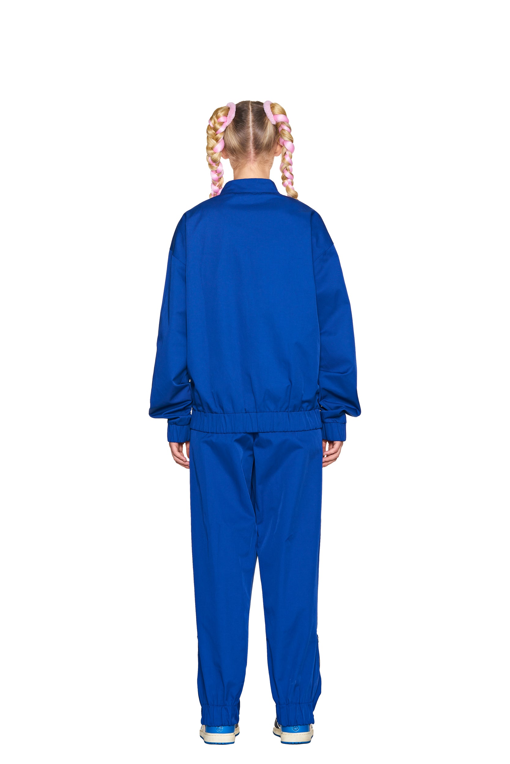 A person with pink and blonde braided hair faces away, wearing 6PM TRACK PANTS BLUE with a loose fit and sneakers, set against a white background.