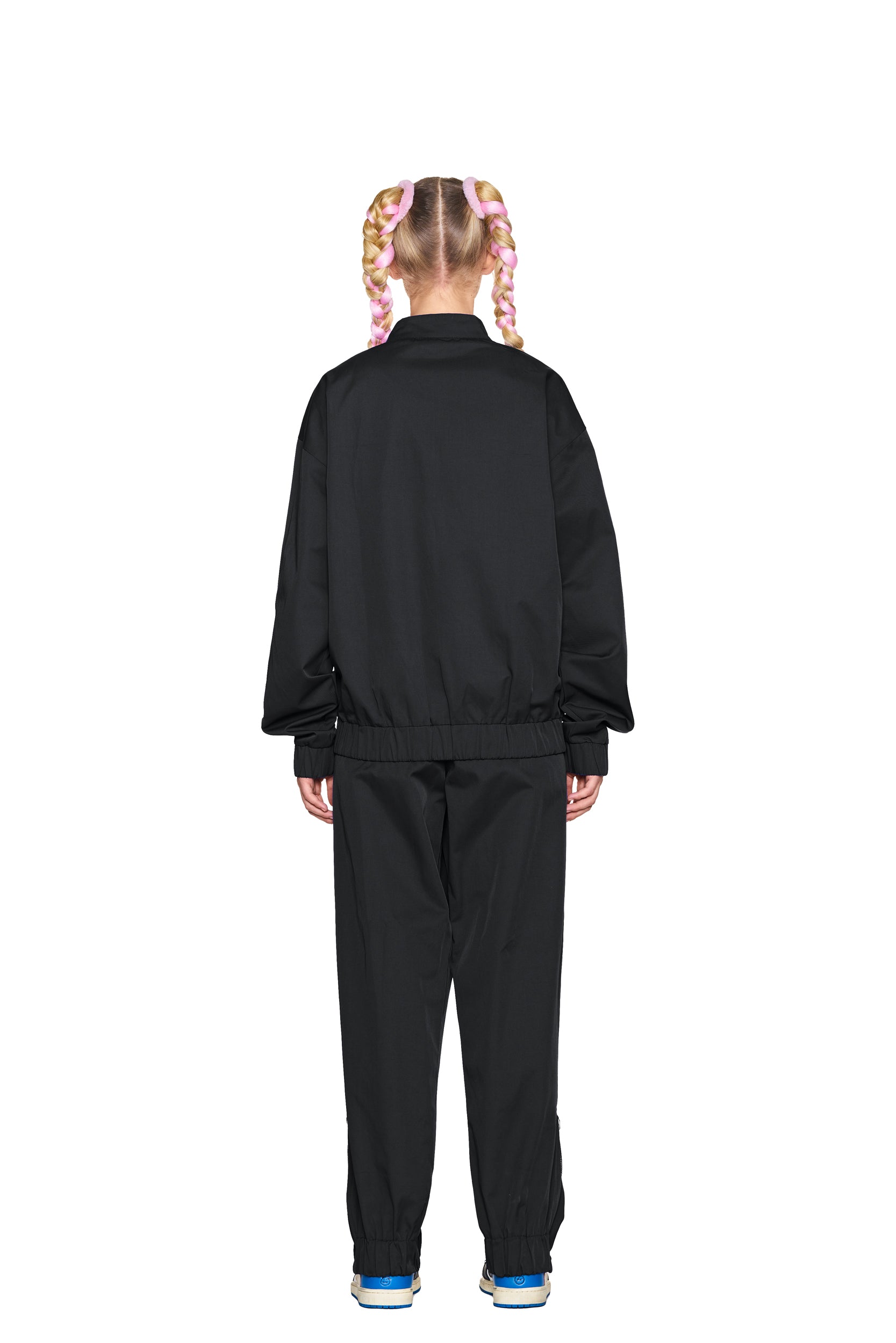 A person with two blonde braids with pink highlights faces away, wearing a black jacket and 6PM TRACK PANTS BLACK paired with blue and white sneakers, against a plain white background.