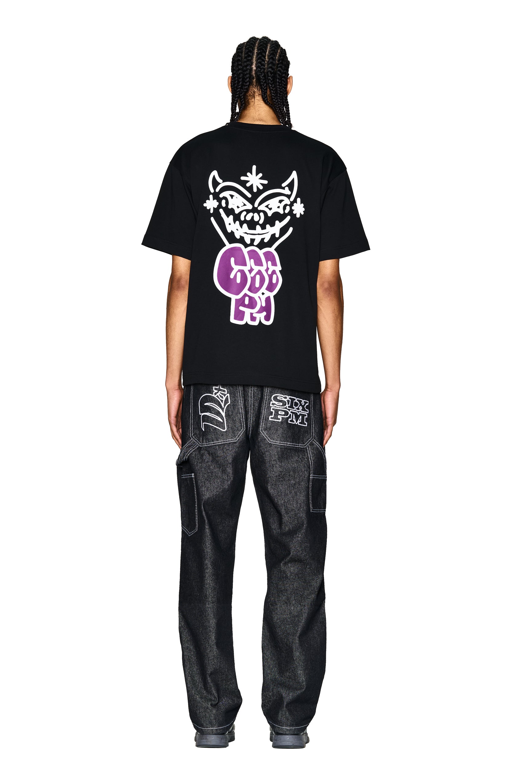 A person with braided hair, seen from behind, wears the 6PM LOGO T-SHIRT BLACK, which features a large white and purple grinning cartoon face and bold lettering, paired with loose dark jeans with stitched designs on the back pockets.