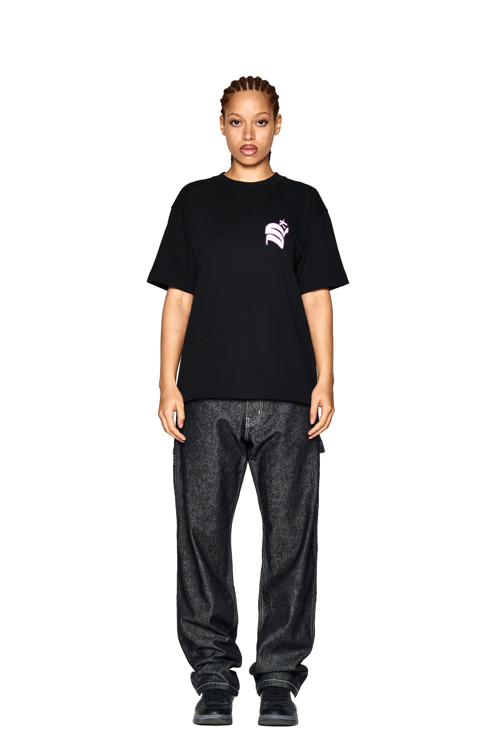 A person faces forward against a white background, wearing the 6PM LOGO T-SHIRT BLACK featuring a small pink chest graphic, paired with loose dark jeans and black shoes.