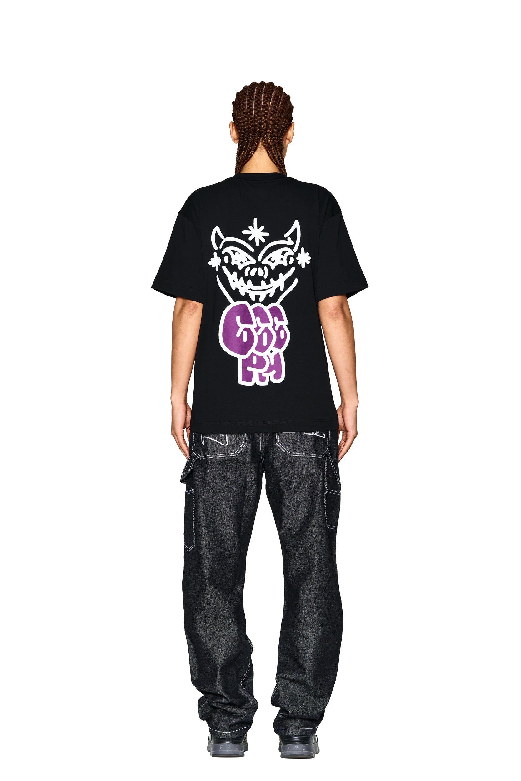 A person stands facing away in a 6PM LOGO T-SHIRT BLACK featuring a large white cartoon face and "GOOD BOY" in purple on the back, paired with dark baggy jeans and sneakers, against a white background.
