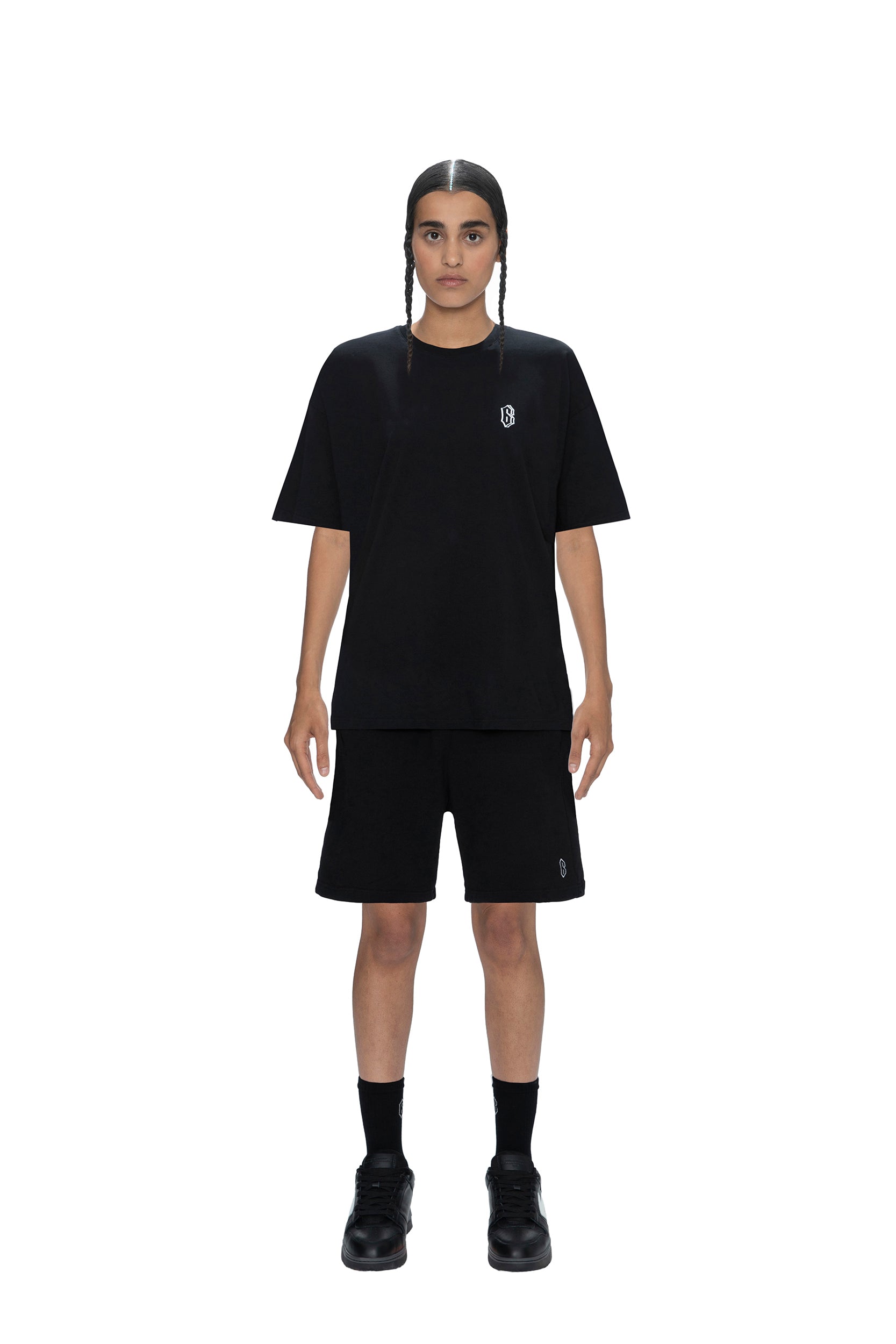 A person with long dark hair in two braids faces forward, wearing the 6PM SIGNATURE T-SHIRT BLACK along with black shorts, socks, and shoes, photographed against a white background.