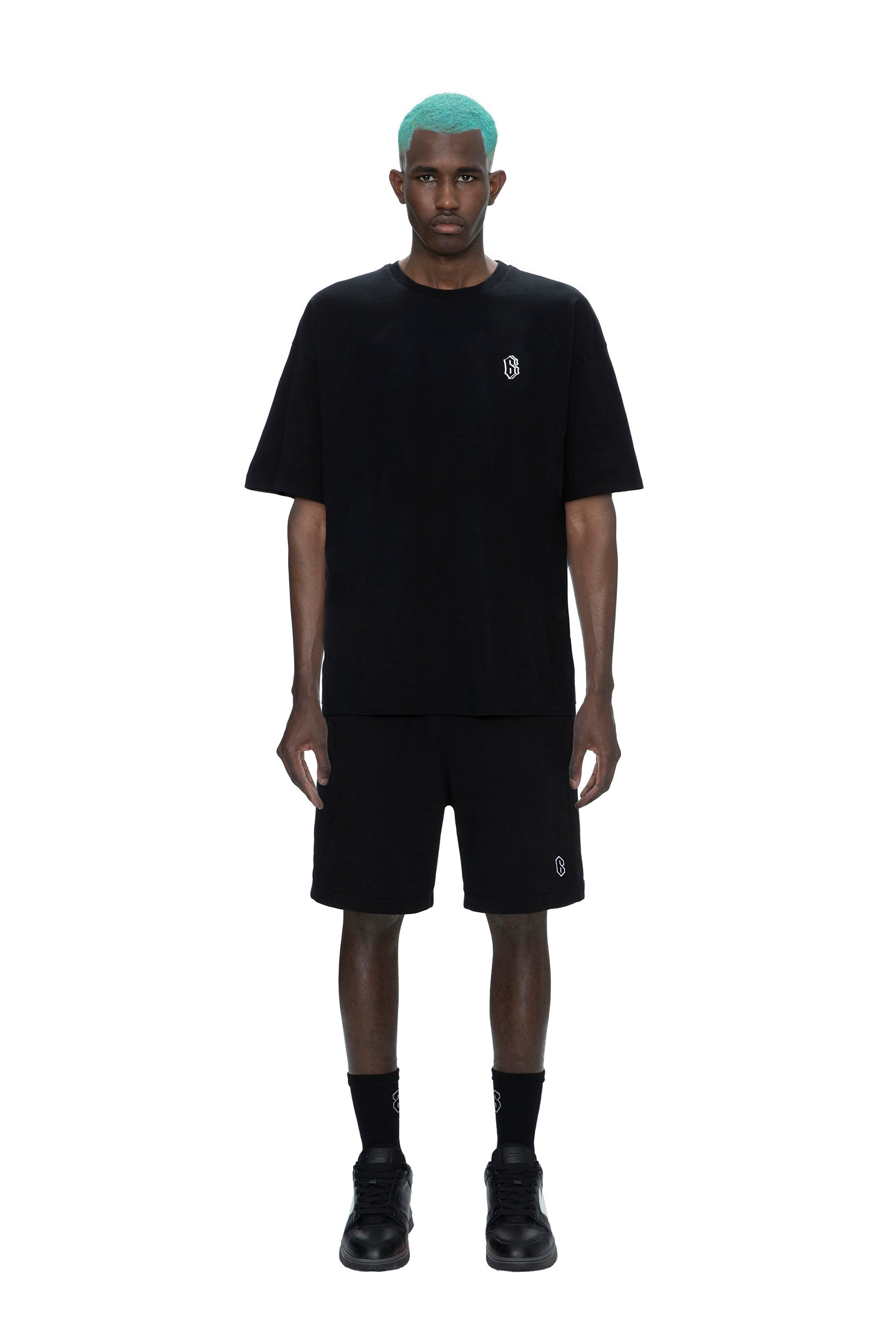 A man with teal hair wears the 6PM SIGNATURE T-SHIRT BLACK, black shorts—both featuring a small white logo—plus black socks and black shoes, standing against a plain white background.