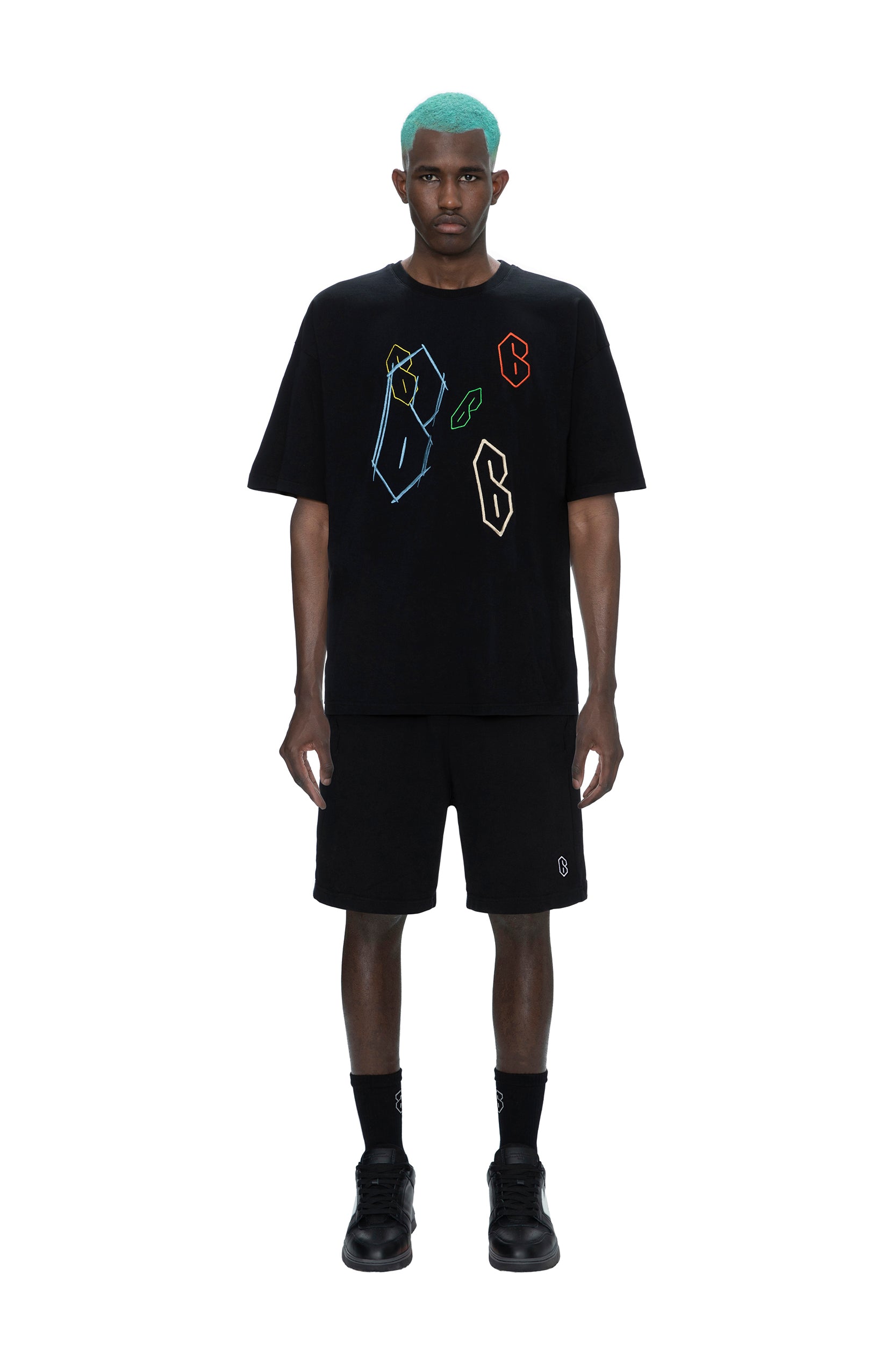A man with teal hair stands facing forward, wearing the 6PM SUPER 6 T-SHIRT BLACK, black shorts, black socks, and black sneakers against a plain white background.