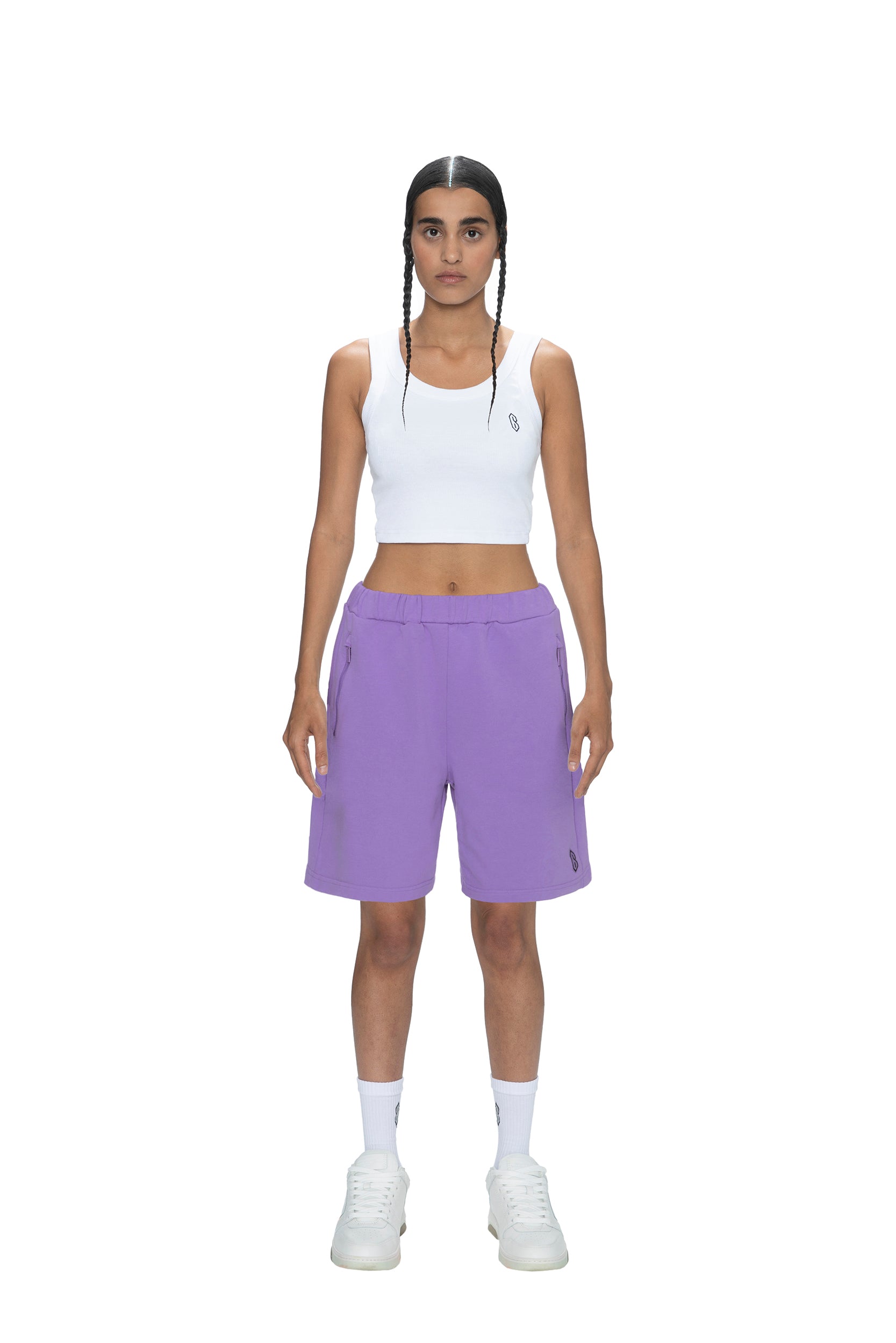 A person faces forward against a plain white background, wearing 6PM’s SWEATSHORTS LILAC, a white crop top, white calf-high socks, and white sneakers. Their hair is styled in two long braids.