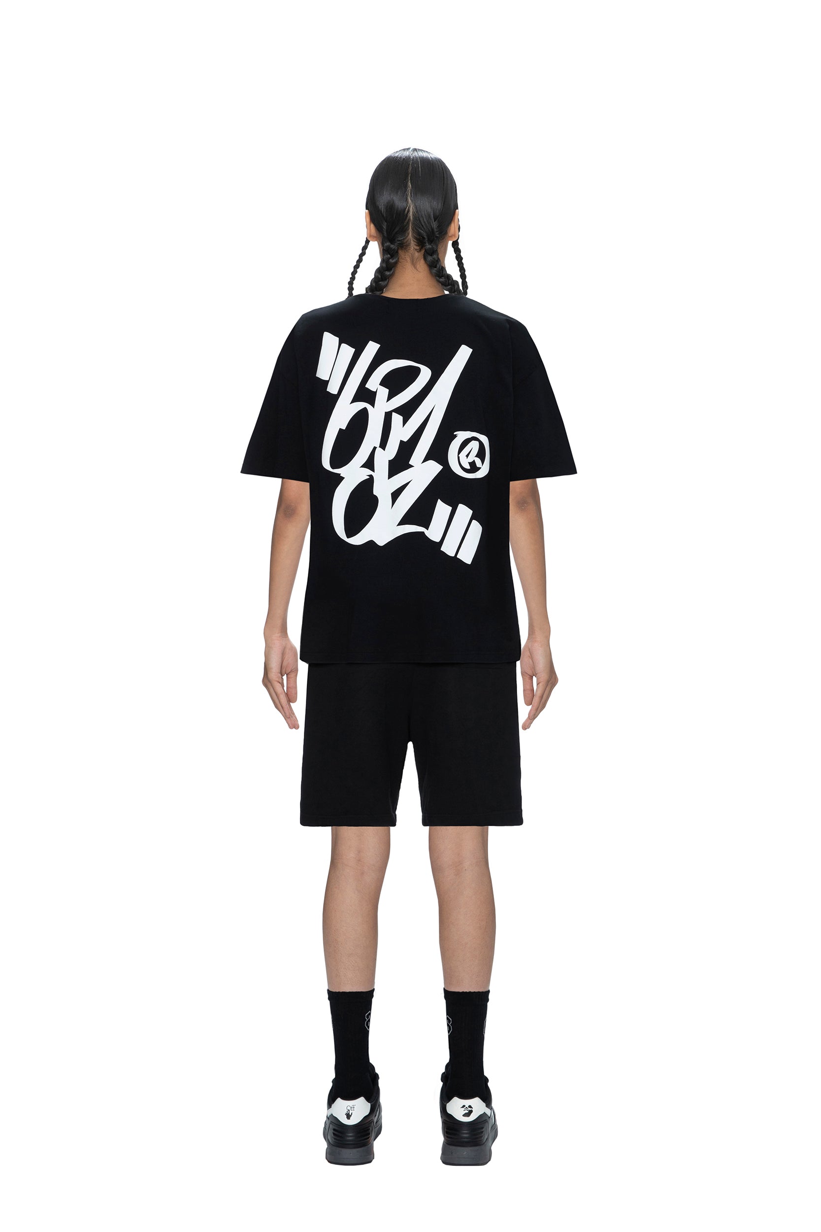 A person with braided hair stands facing away, wearing the 6PM SIGNATURE T-SHIRT BLACK featuring bold white graffiti-style text on the back, paired with black shorts, socks, and sneakers, set against a plain white background.