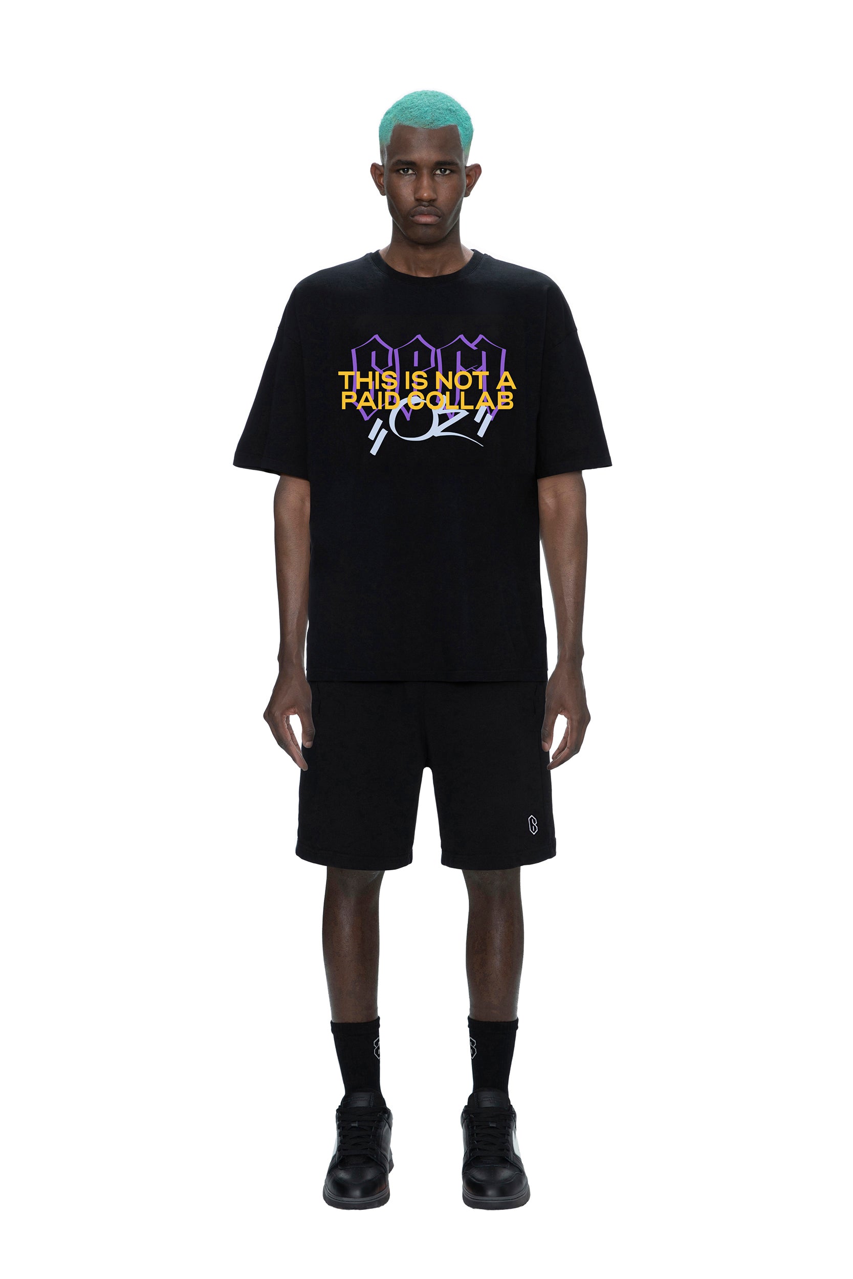 A man with turquoise hair faces forward in a loose black 6PM PAID COLLAB T-SHIRT with bold yellow and white text, paired with black shorts, socks, and shoes, all set against a white background.