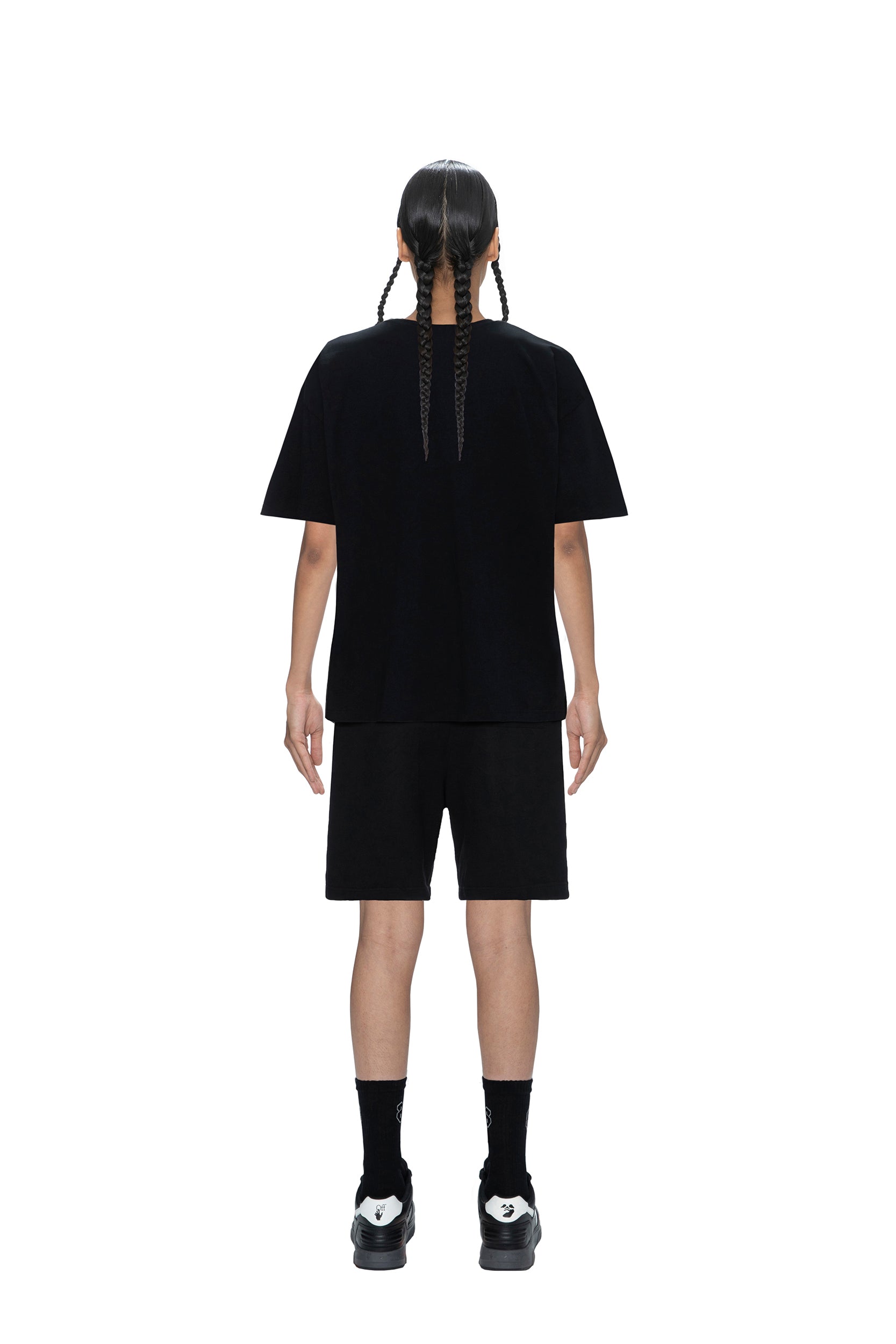 A person with long, braided hair faces away, wearing the 6PM PAID COLLAB T-SHIRT BLACK, black shorts, black socks, and black sneakers, standing against a plain white background.