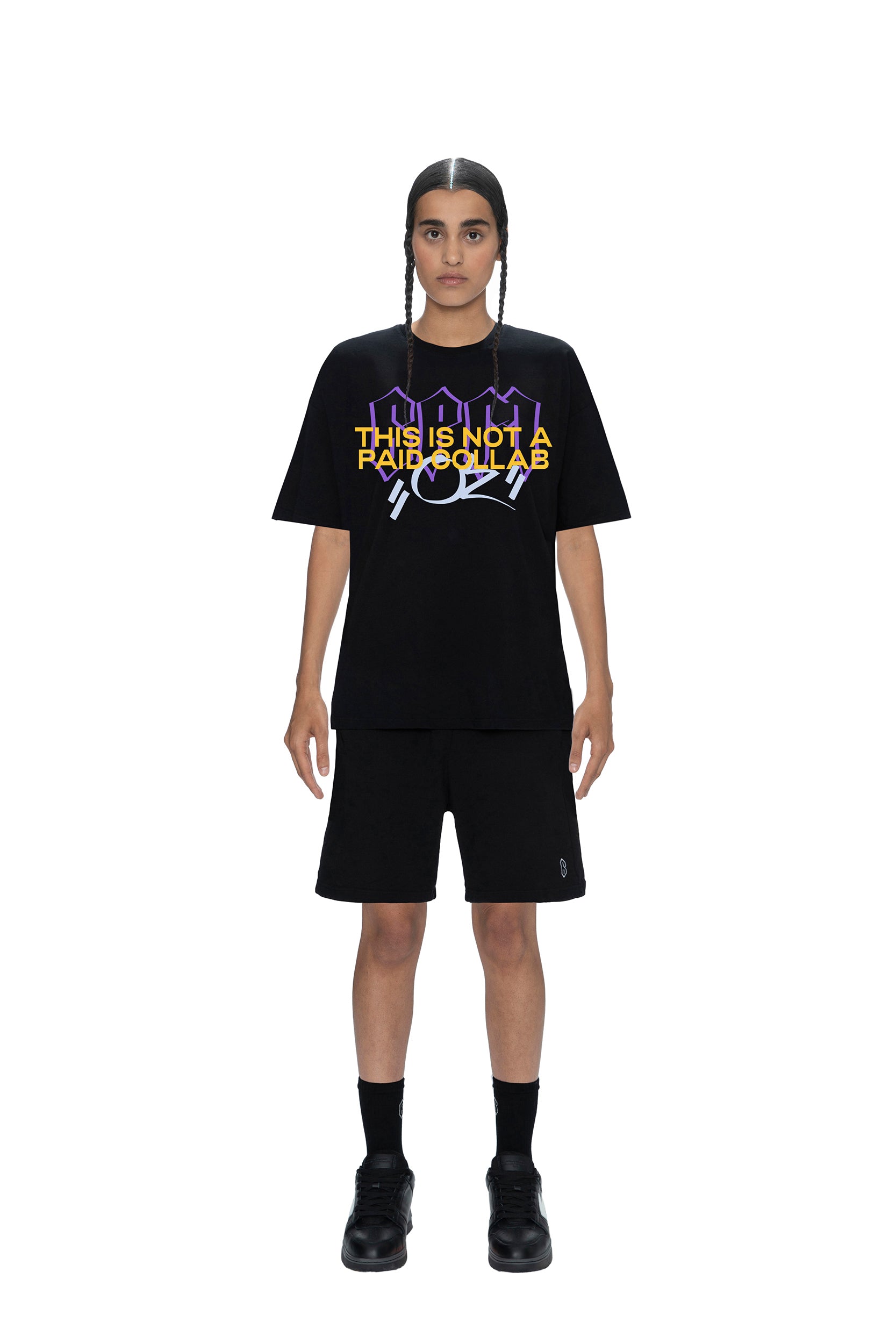 A person with braided hair faces forward, wearing the 6PM PAID COLLAB T-SHIRT BLACK featuring purple, yellow, and white text, paired with black shorts, socks, and shoes, all set against a white background.