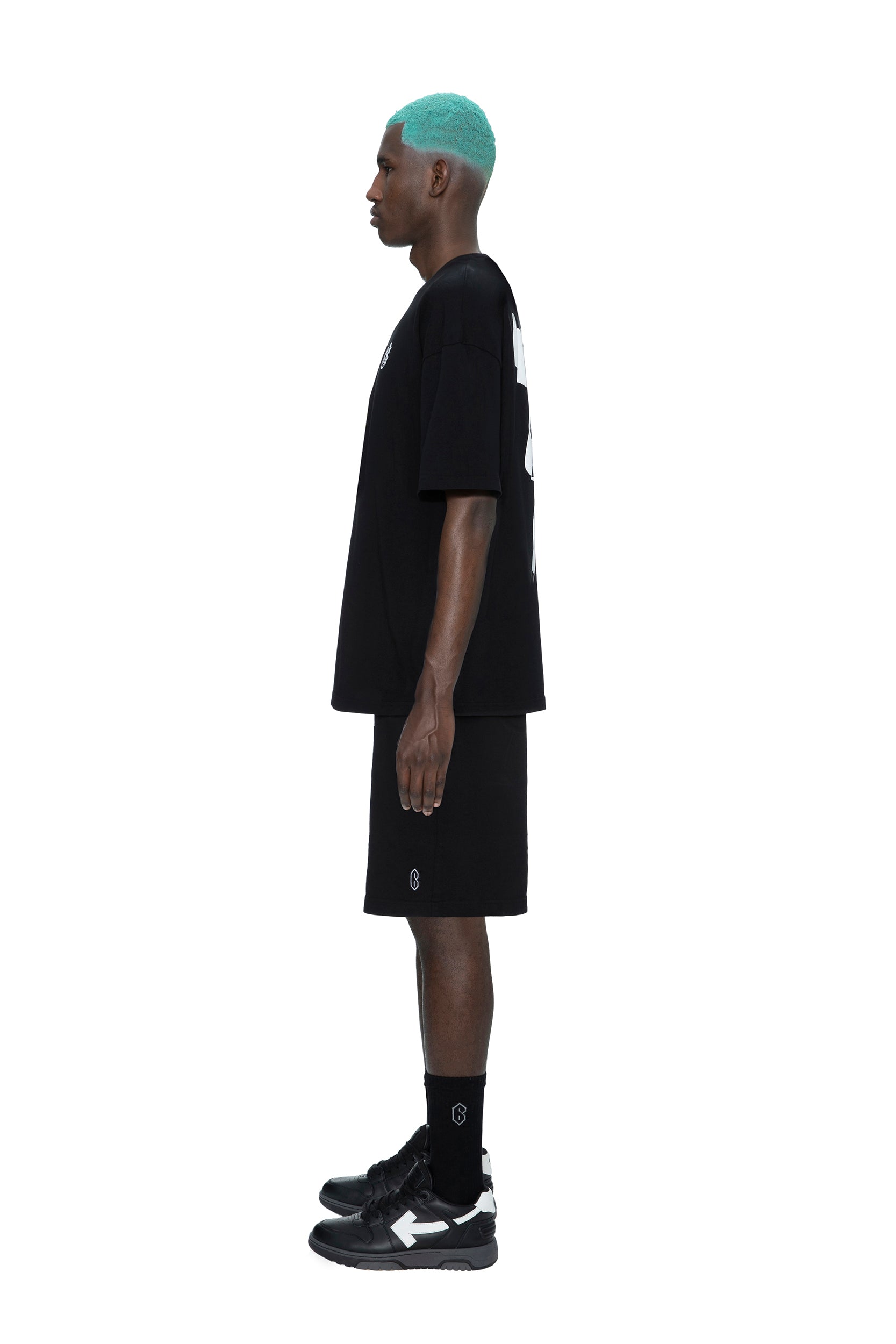 A man with teal hair stands in profile against a white background, wearing the 6PM SIGNATURE T-SHIRT BLACK, black shorts, black socks, and black sneakers with white soles.