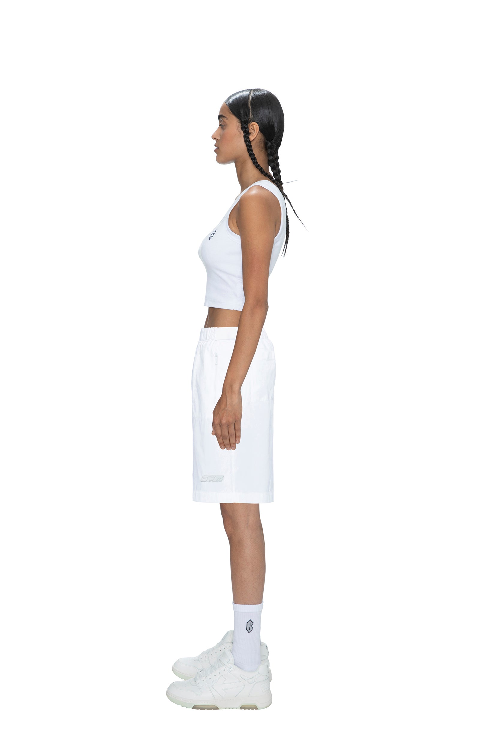 A person with long braids stands in profile facing left, wearing 6PM TECH SHORTS WHITE, a white sleeveless crop top, white socks, and white sneakers against a plain white background.