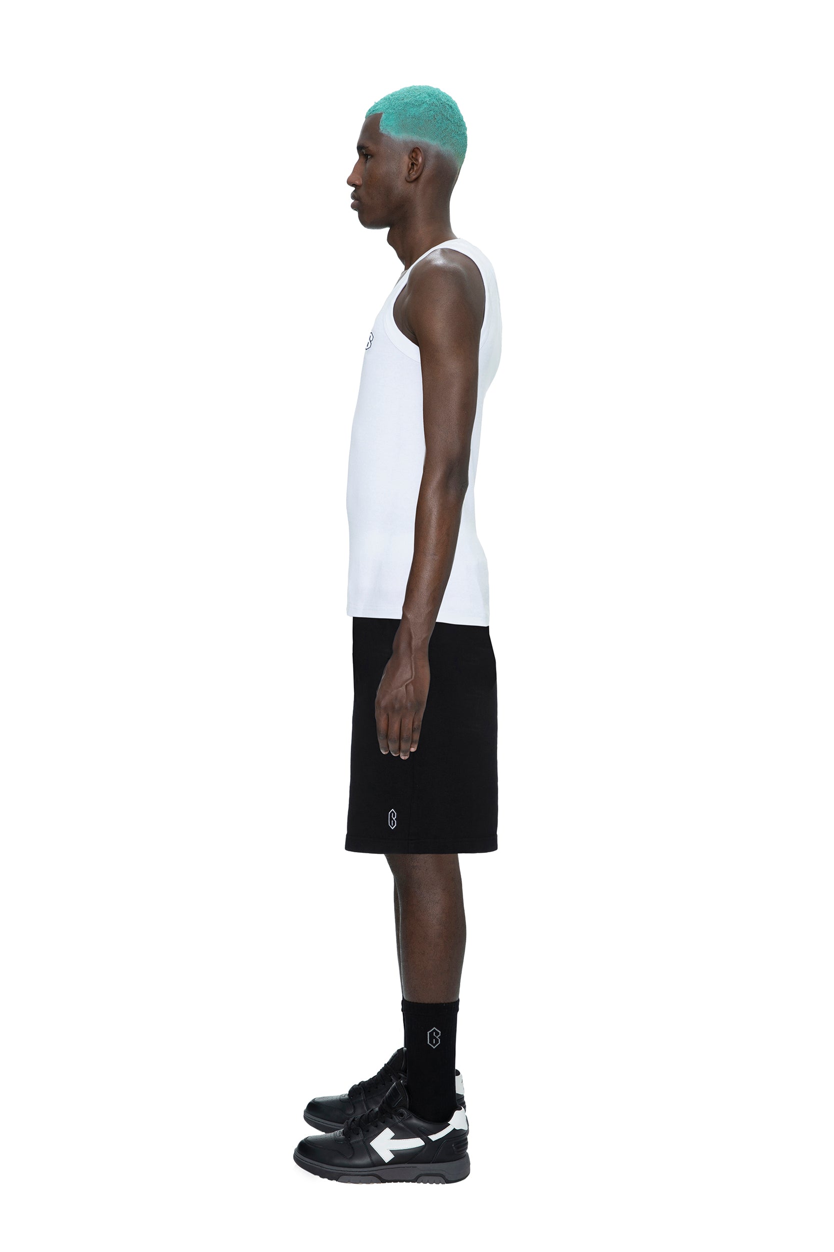 A man with teal hair wears a white tank top, 6PM SWEATSHORTS BLACK, black crew socks, and black sneakers, standing in profile against a plain white background.