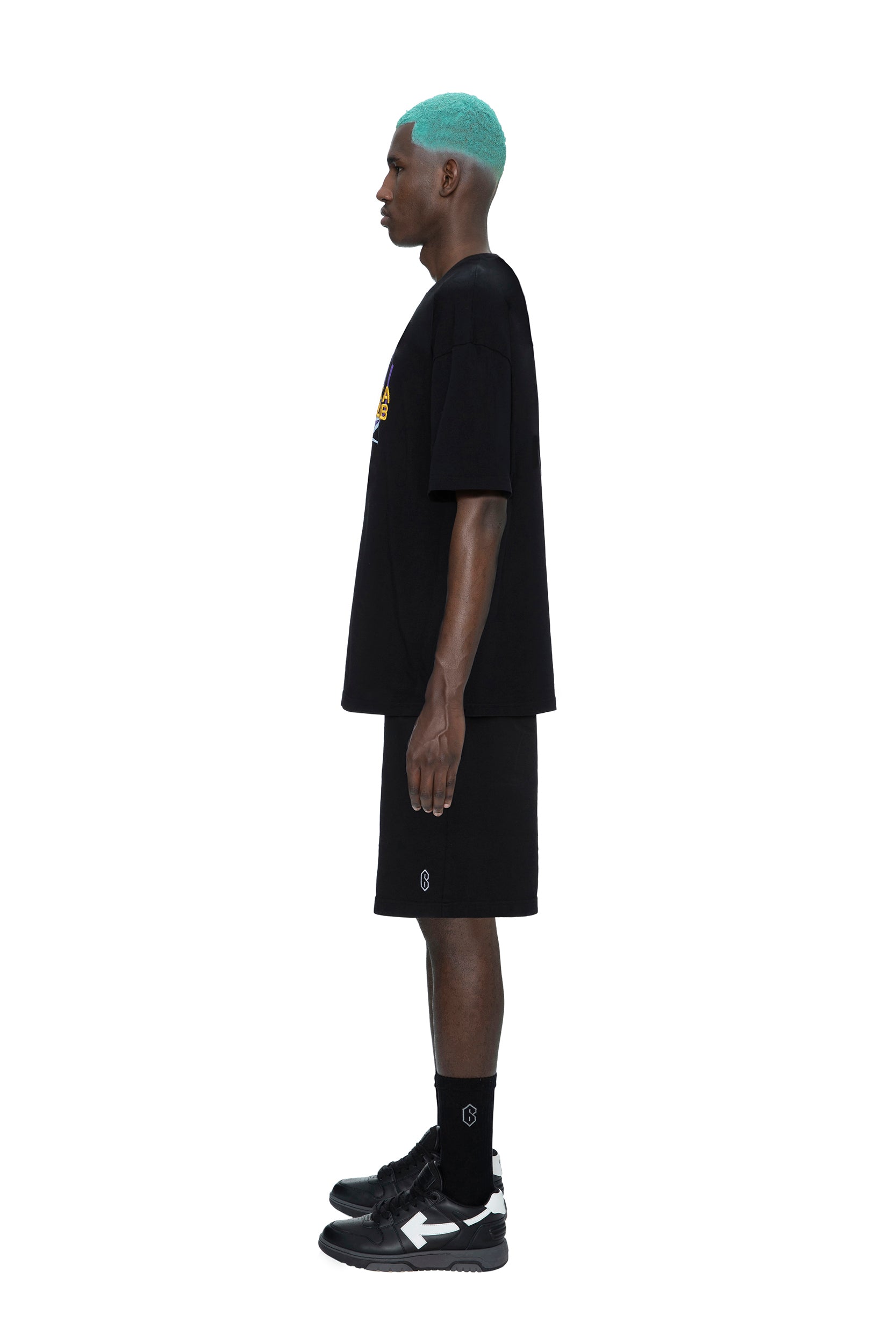 A man with teal hair stands in profile against a white background, wearing the 6PM PAID COLLAB T-SHIRT BLACK, paired with black shorts, black socks, and black sneakers.